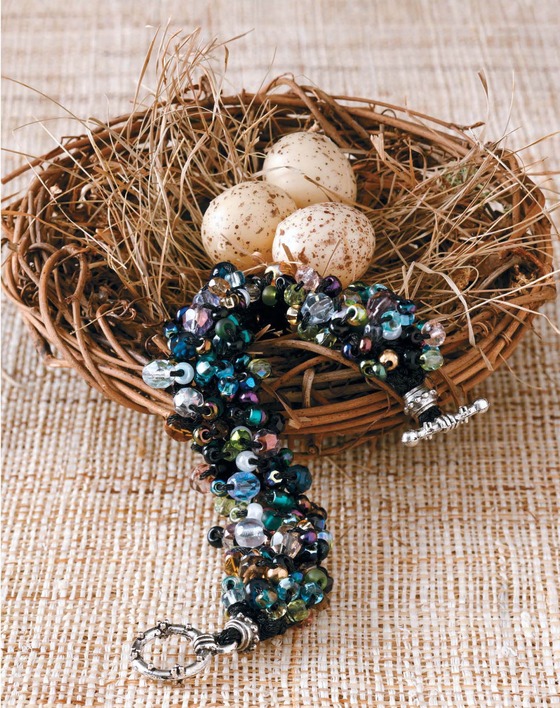 On a straw mat, a woven bird’s nest with three speckled eggs sits behind a beaded bracelet from Leisure Arts' "Beaded Bracelets to Knit," featuring shiny blue, green, and black beads with a silver clasp—great for beginner jewelry makers.