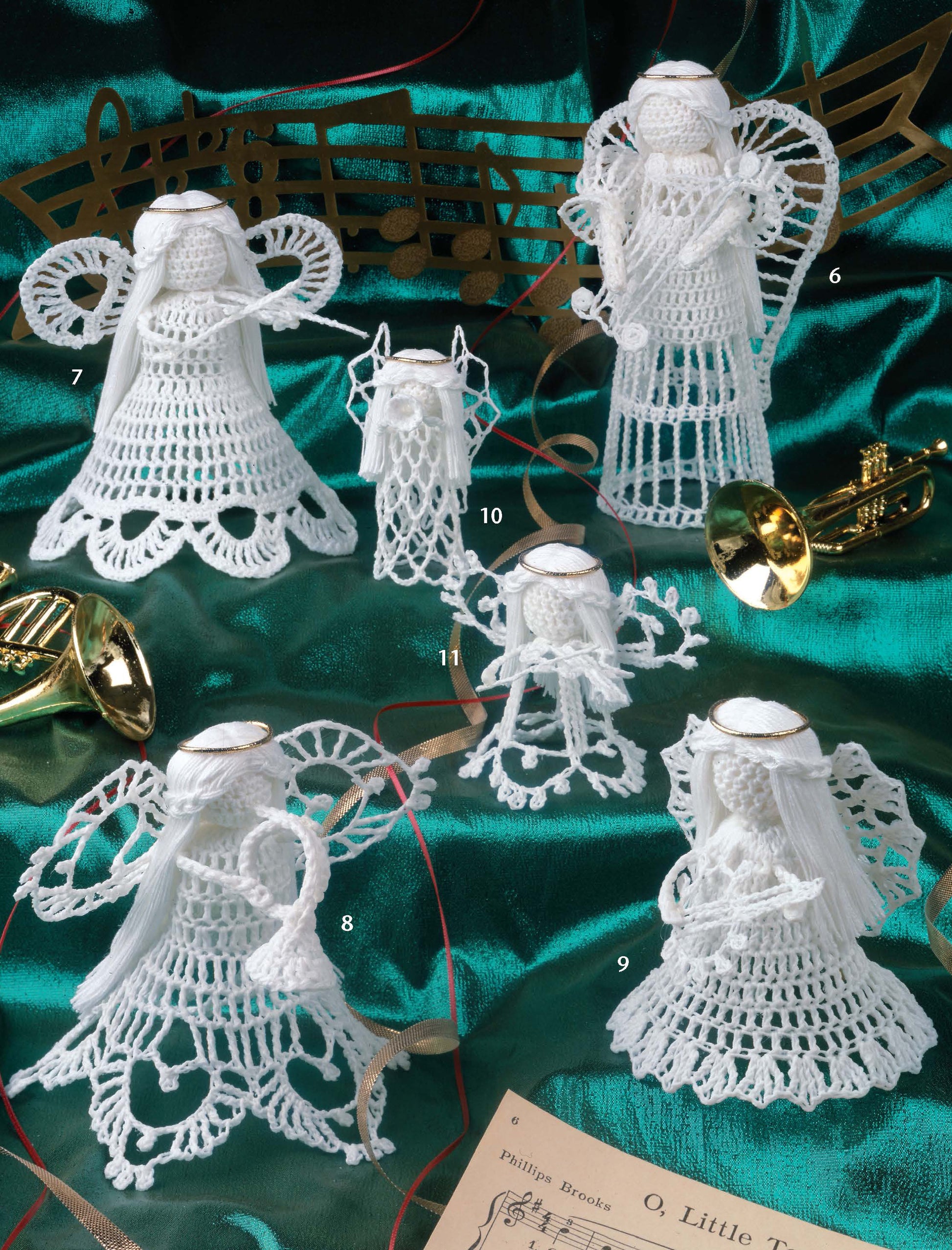 Five delicate lace-weight crocheted angel figurines and a nativity lamb, as featured in the "Big Book of Thread Ornaments" by Leisure Arts, are displayed on green satin with gold musical notes, mini instruments, and sheet music.
