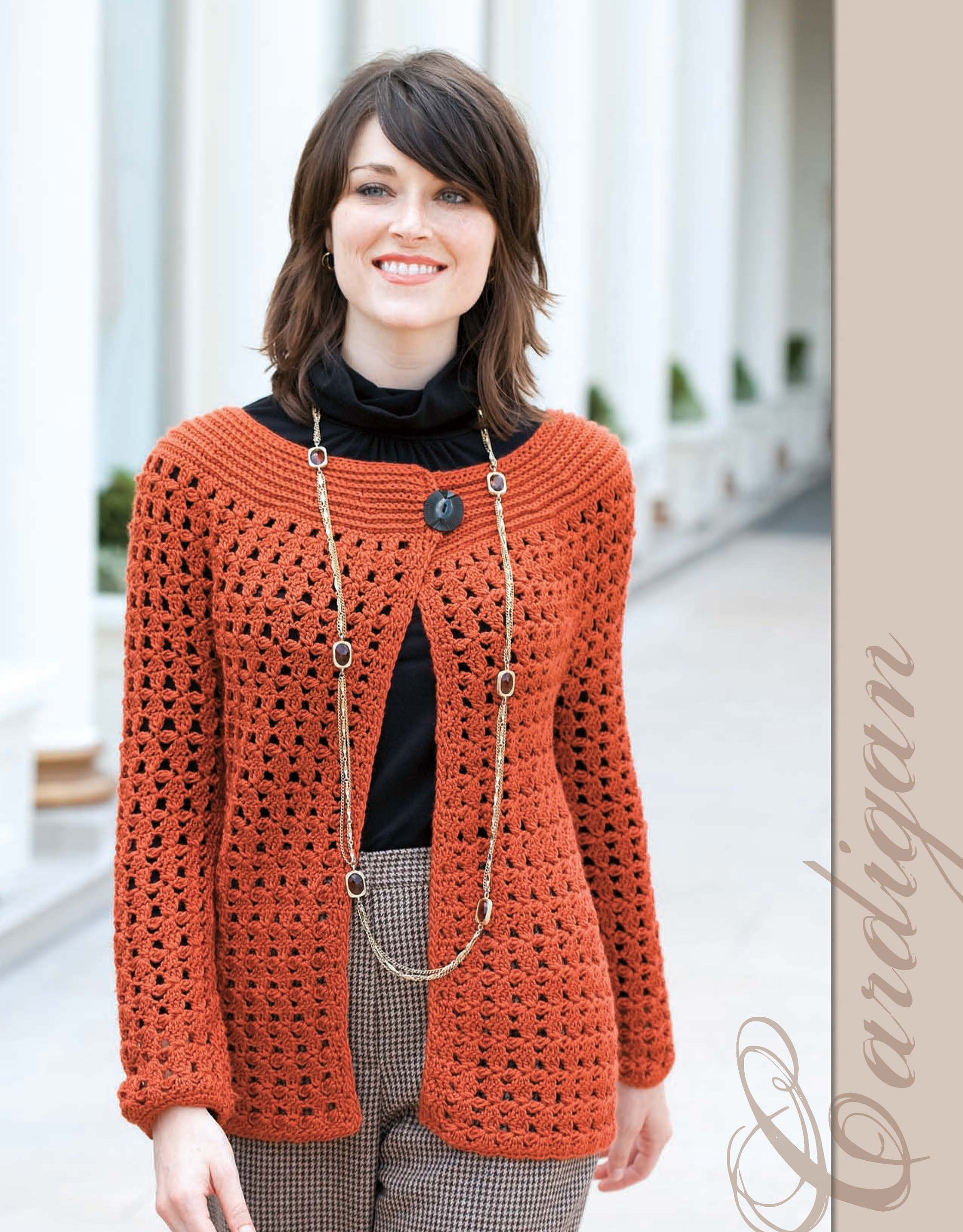 A woman with short brown hair smiles in a burnt orange cardigan from The Crochet Closet by Leisure Arts over a black top and checkered pants, standing in a sunny outdoor corridor. "Cardigan" appears vertically on the right.