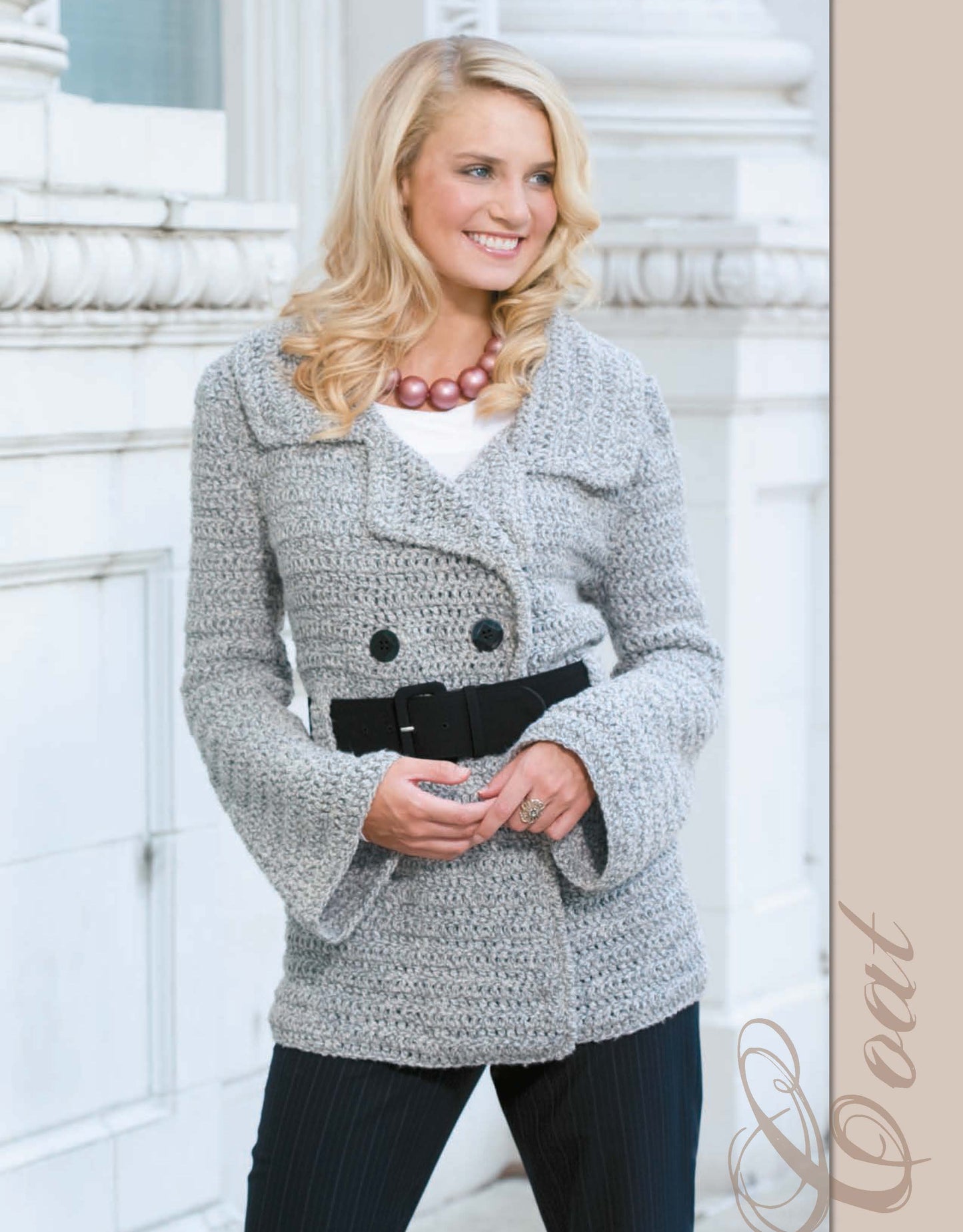 A smiling blonde woman models a chunky light gray knit coat from The Crochet Closet by Leisure Arts, styled with a black belt, black pants, and a pink beaded necklace, standing before a white architectural background.