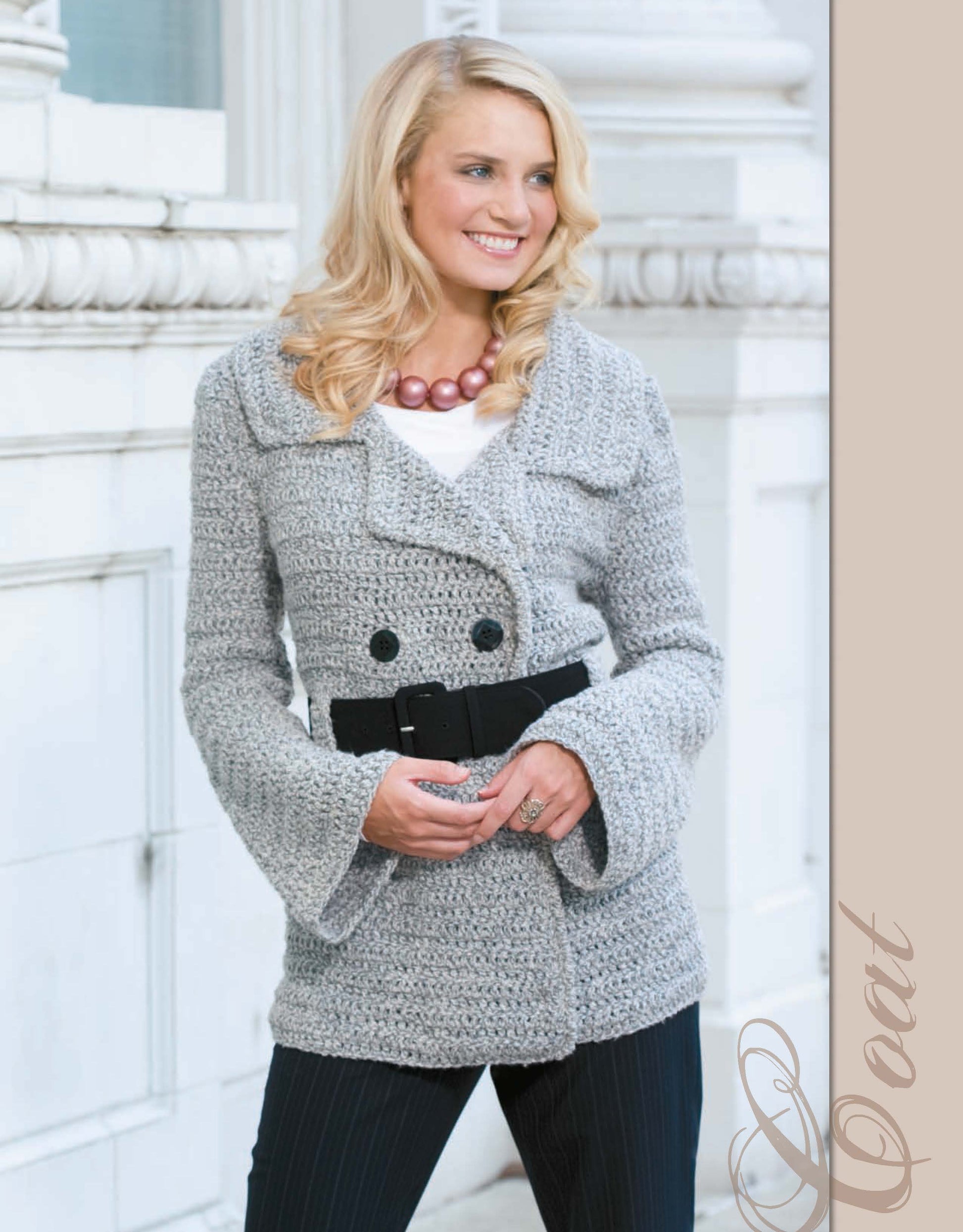 A smiling blonde woman models a chunky light gray knit coat from The Crochet Closet by Leisure Arts, styled with a black belt, black pants, and a pink beaded necklace, standing before a white architectural background.