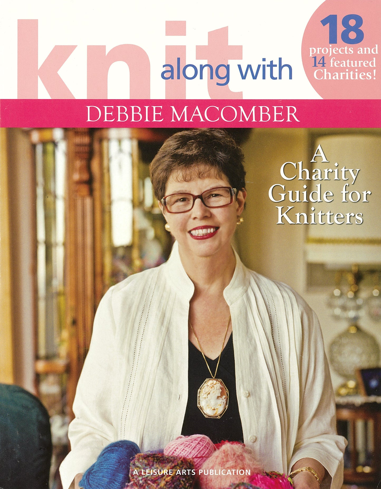 A smiling woman with short dark hair and glasses stands before a table of colorful yarn. The cover reads: Knit Along with Debbie Macomber—A Charity Guide for Knitters by Leisure Arts, featuring 18 projects for 14 charities, ideal for knitters and beginners.