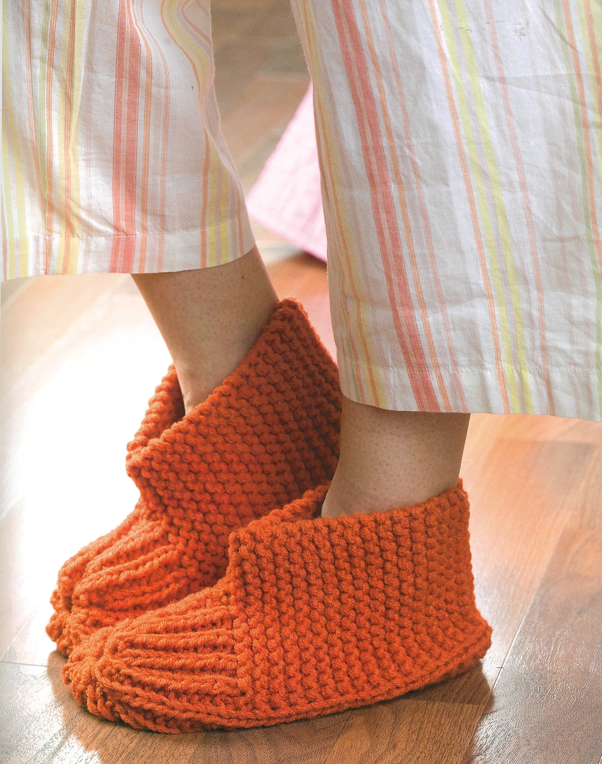 A person in loose, striped pajama pants and bright orange knitted slippers—ideal for beginner projects from "Knit Along with Debbie Macomber—A Charity Guide for Knitters" by Leisure Arts—stands on a wooden floor.