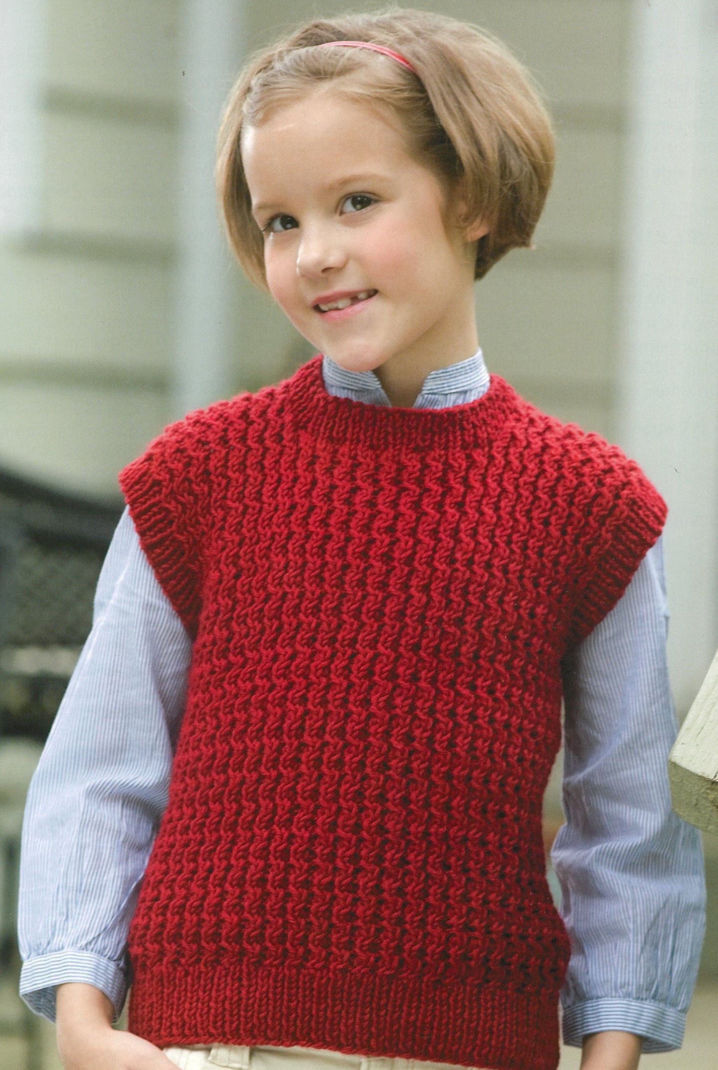 A young child with short brown hair smiles in front of a house, modeling a red sleeveless sweater over a blue shirt—a great starter project for knitters inspired by Leisure Arts' Knit Along with Debbie Macomber: A Charity Guide for Knitters.