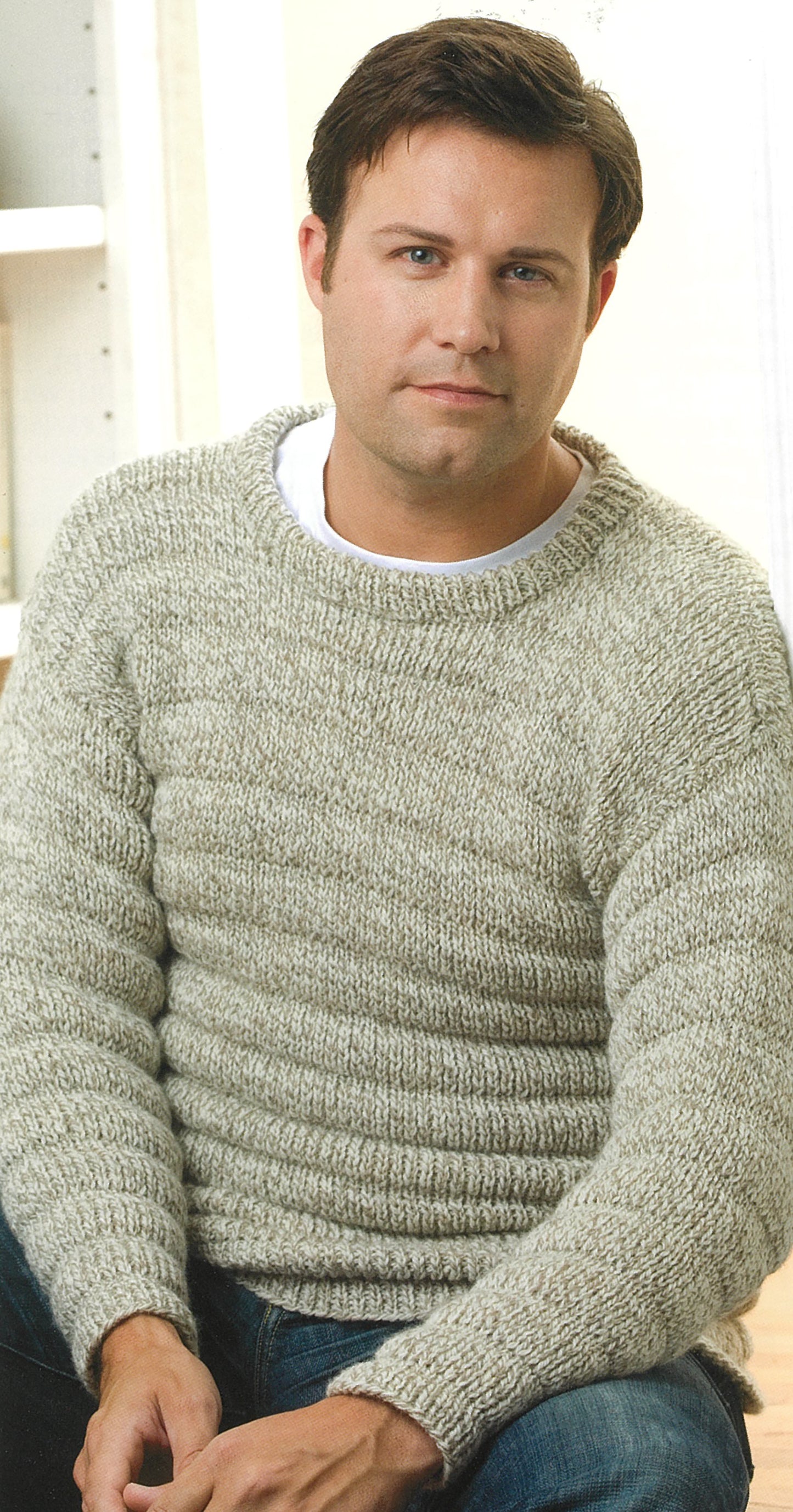 A man with short brown hair in a light gray knitted sweater and jeans sits indoors, facing the camera—ideal inspiration for Leisure Arts’ “Knit Along with Debbie Macomber—A Charity Guide for Knitters.”.