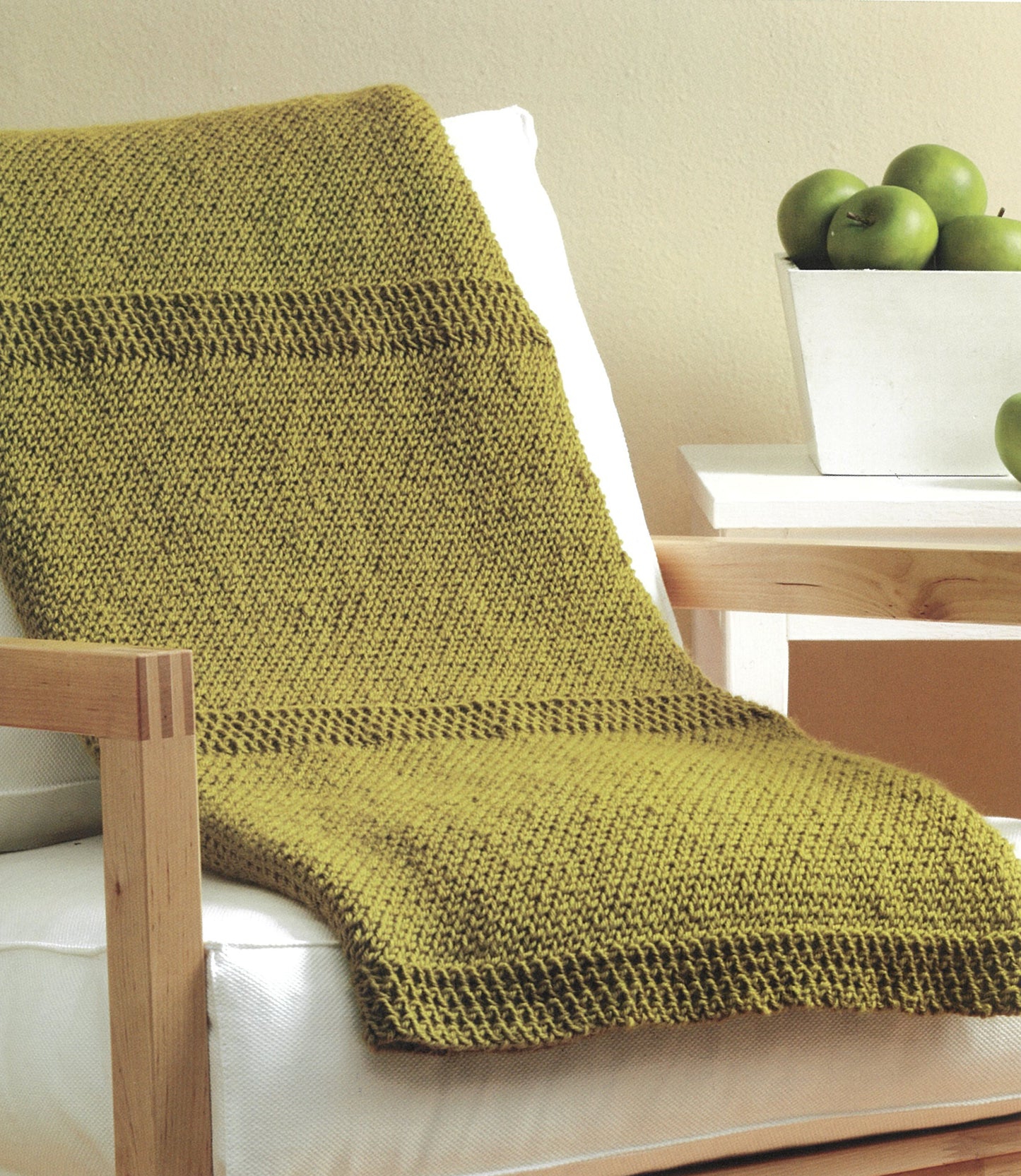 A cozy green blanket from "Knit Along with Debbie Macomber—A Charity Guide for Knitters" by Leisure Arts is draped over a white-cushioned wooden chair next to a bowl of green apples on a wooden table.