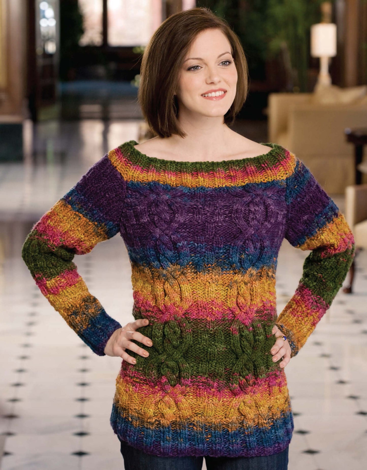 A woman stands indoors, smiling and wearing a colorful, long-sleeve knit sweater with horizontal stripes from the Deborah Newton's Cable Collection by Leisure Arts. The background features a polished floor and blurred furniture.