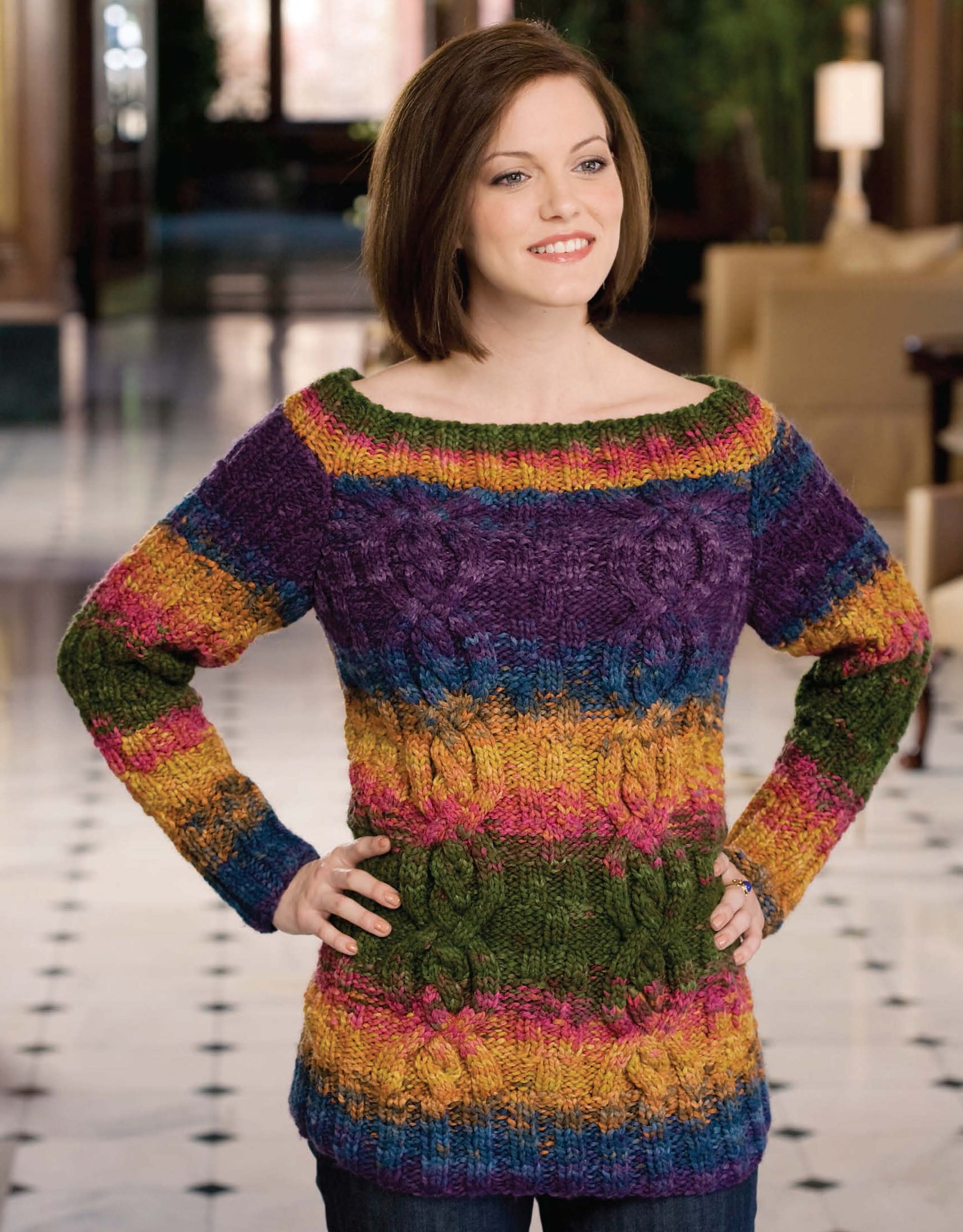 A woman stands indoors, smiling and wearing a colorful, long-sleeve knit sweater with horizontal stripes from the Deborah Newton's Cable Collection by Leisure Arts. The background features a polished floor and blurred furniture.