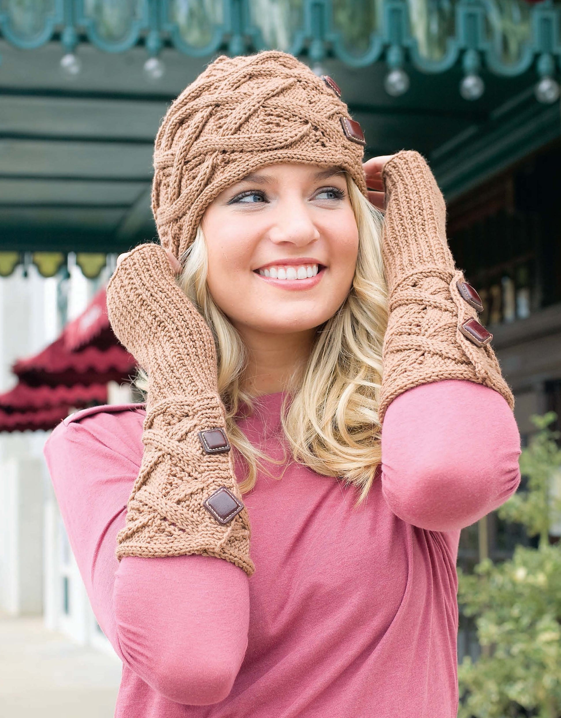 A smiling woman in cozy accessories from Leisure Arts’ Deborah Newton’s Cable Collection stands outdoors, showcasing a pink top, tan knit hat, and fingerless mittens that perfectly pair with cabled cardigans and women’s sweaters.
