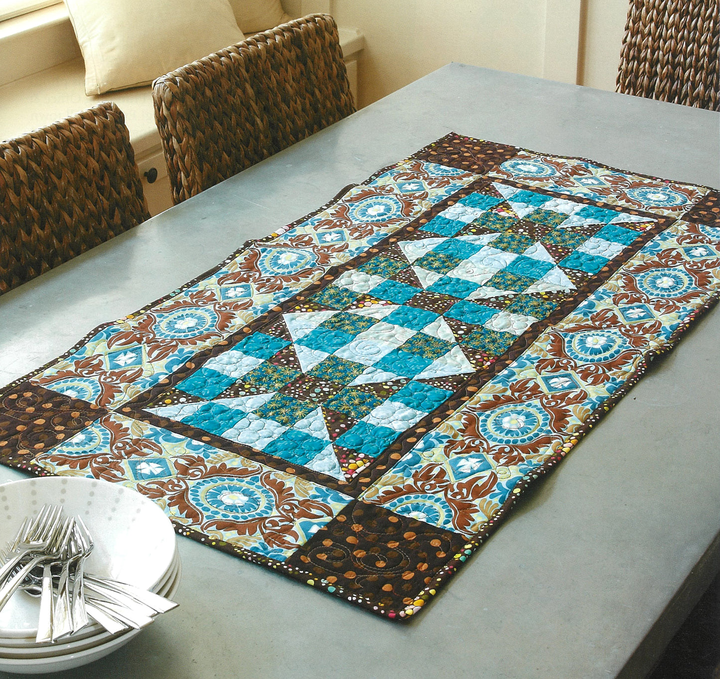 A vibrant quilted table runner made with fabrics inspired by Pat Sloan's Camera, Color, Quilts! from Leisure Arts adds color to a light gray dining table, surrounded by woven chairs and a stack of white plates with silverware.