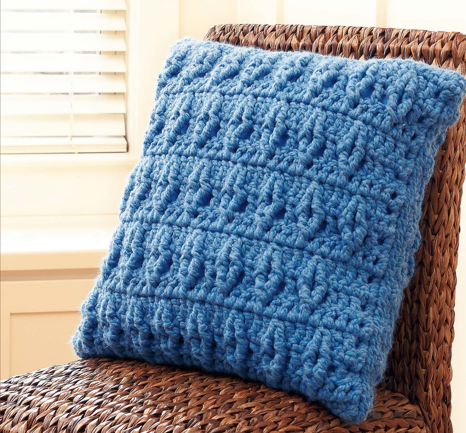 The Quick Comforts Home Makeover pillow by Leisure Arts features a chunky blue crochet design with a textured diamond pattern, displayed on a brown wicker chair by a window with white horizontal blinds.