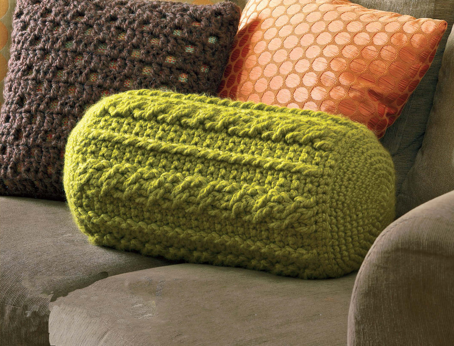 A green, chunky-knit cylindrical pillow from Leisure Arts' Quick Comforts Home Makeover collection sits on a brown couch, with a textured brown pillow and an orange hexagon-patterned pillow behind it.