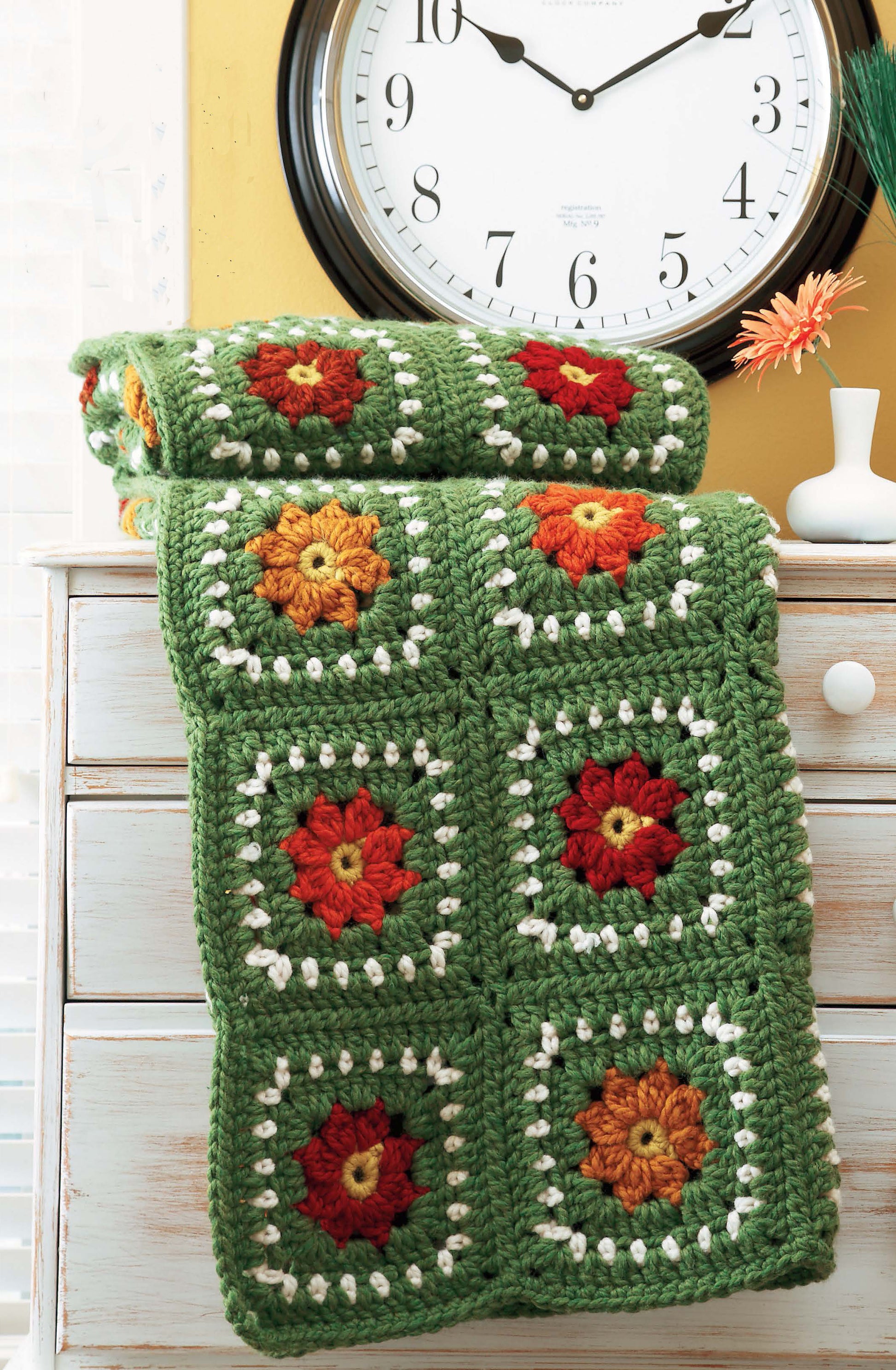 The Leisure Arts Quick Comforts Home Makeover green crocheted blanket with colorful floral granny squares is folded on a white dresser under a wall clock displaying 11:07.