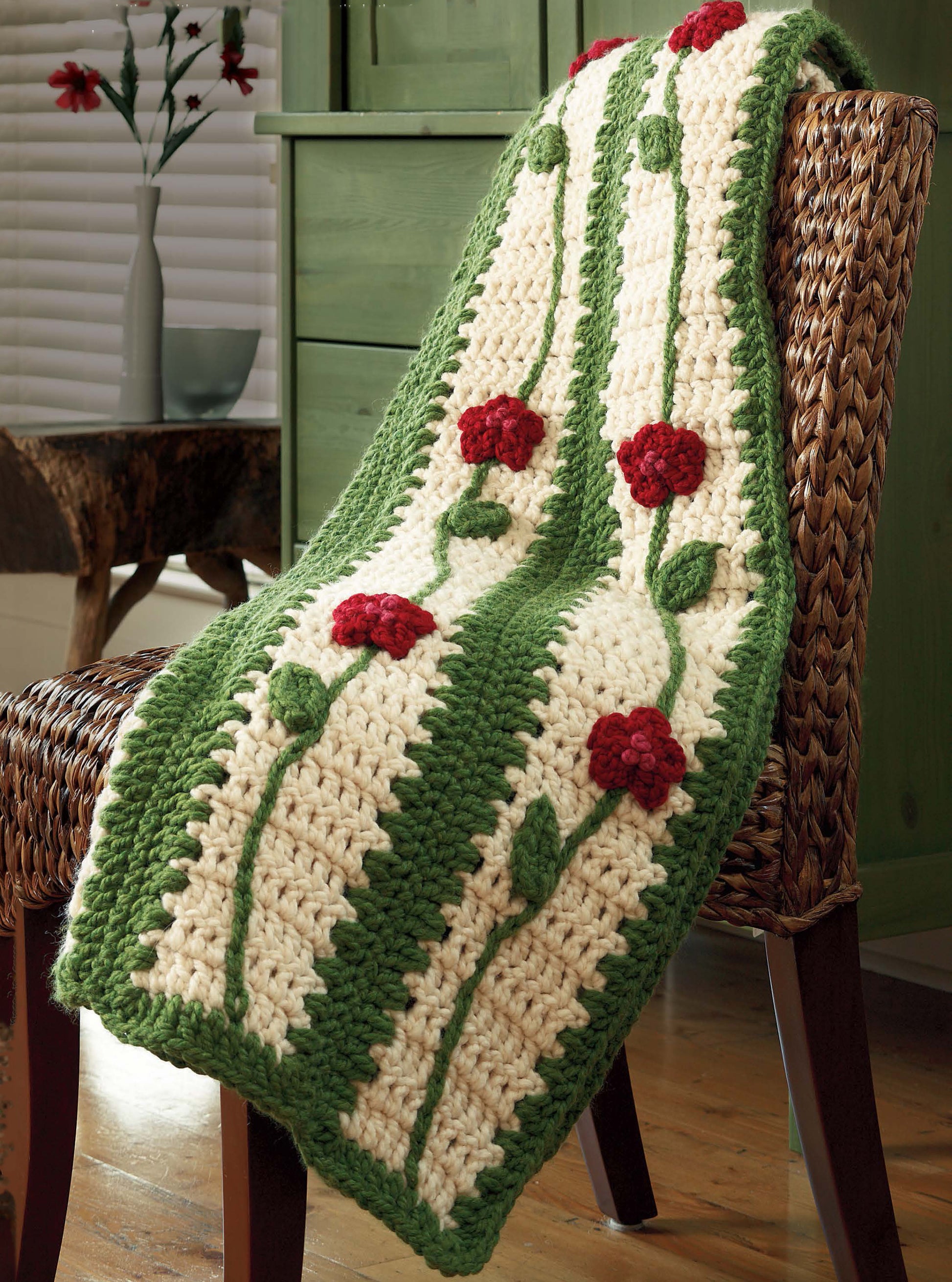A cozy, sunlit room with wooden floors features the "Quick Comforts Home Makeover" crocheted blanket by Leisure Arts, draped over a wicker chair. It has a cream background with green stripes and red flower patterns inspired by classic crochet afghans.