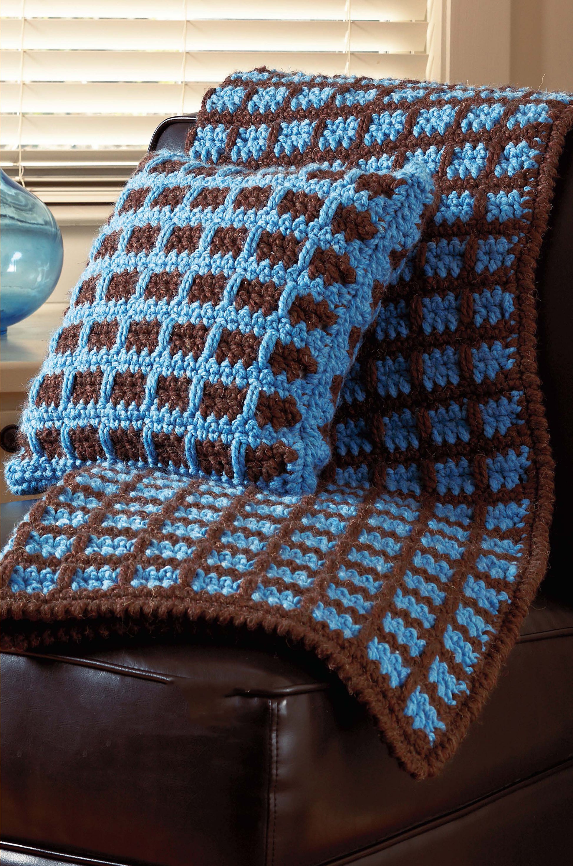 The Quick Comforts Home Makeover set by Leisure Arts features a blue and brown checkered crocheted pillow and blanket, crafted with super bulky yarn and shown on a dark brown leather chair near a window with white blinds.