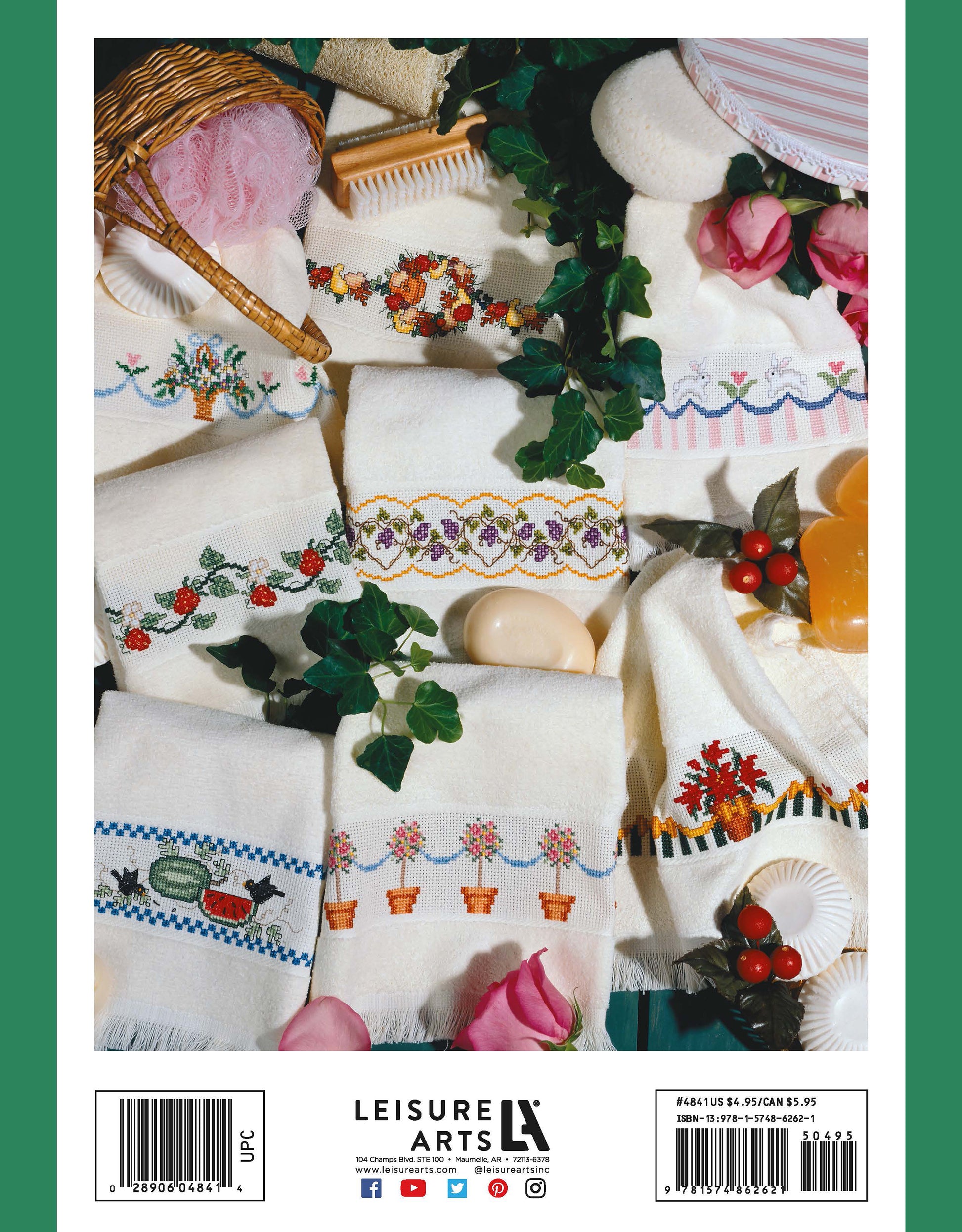 Favorite Fingertips hand towels by Leisure Arts feature colorful embroidered cross stitch designs—flowers, fruit, and geometric patterns—and are displayed with soap, artificial cherries, pink roses, ivy, and a wicker basket on a white surface.