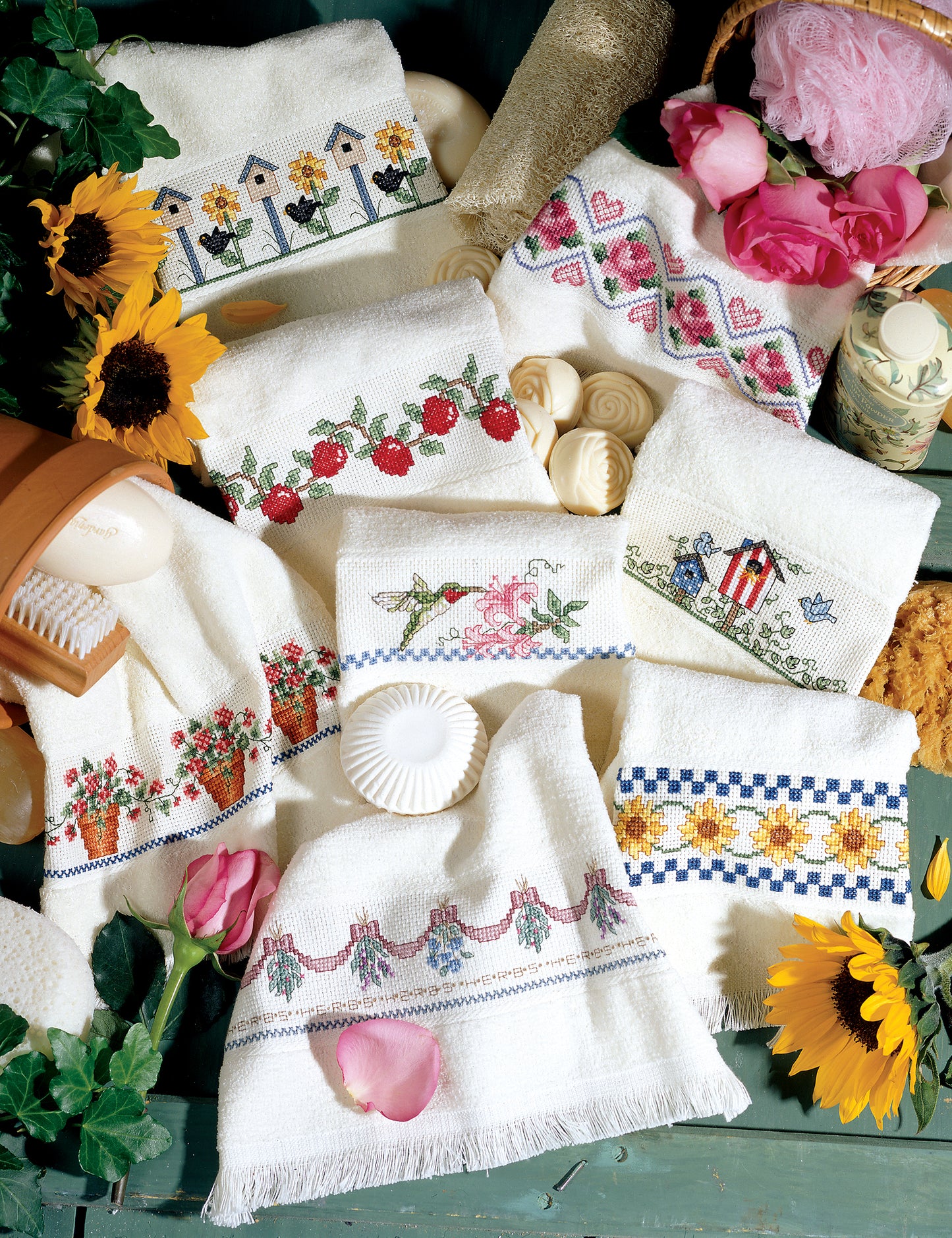 Favorite Fingertips by Leisure Arts features white hand towels with beginner cross stitch designs—flowers, strawberries, hearts, and birds—displayed with sunflowers, rose petals, and bath items on a rustic green surface for charming home decor.