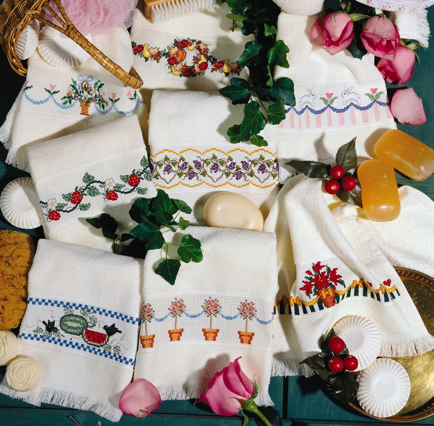 Leisure Arts’ Favorite Fingertips features embroidered white towels with floral and fruit designs—ideal for home decor or beginner cross stitch—displayed among pink roses, green ivy, soaps, and decor on a green surface for a charming look.