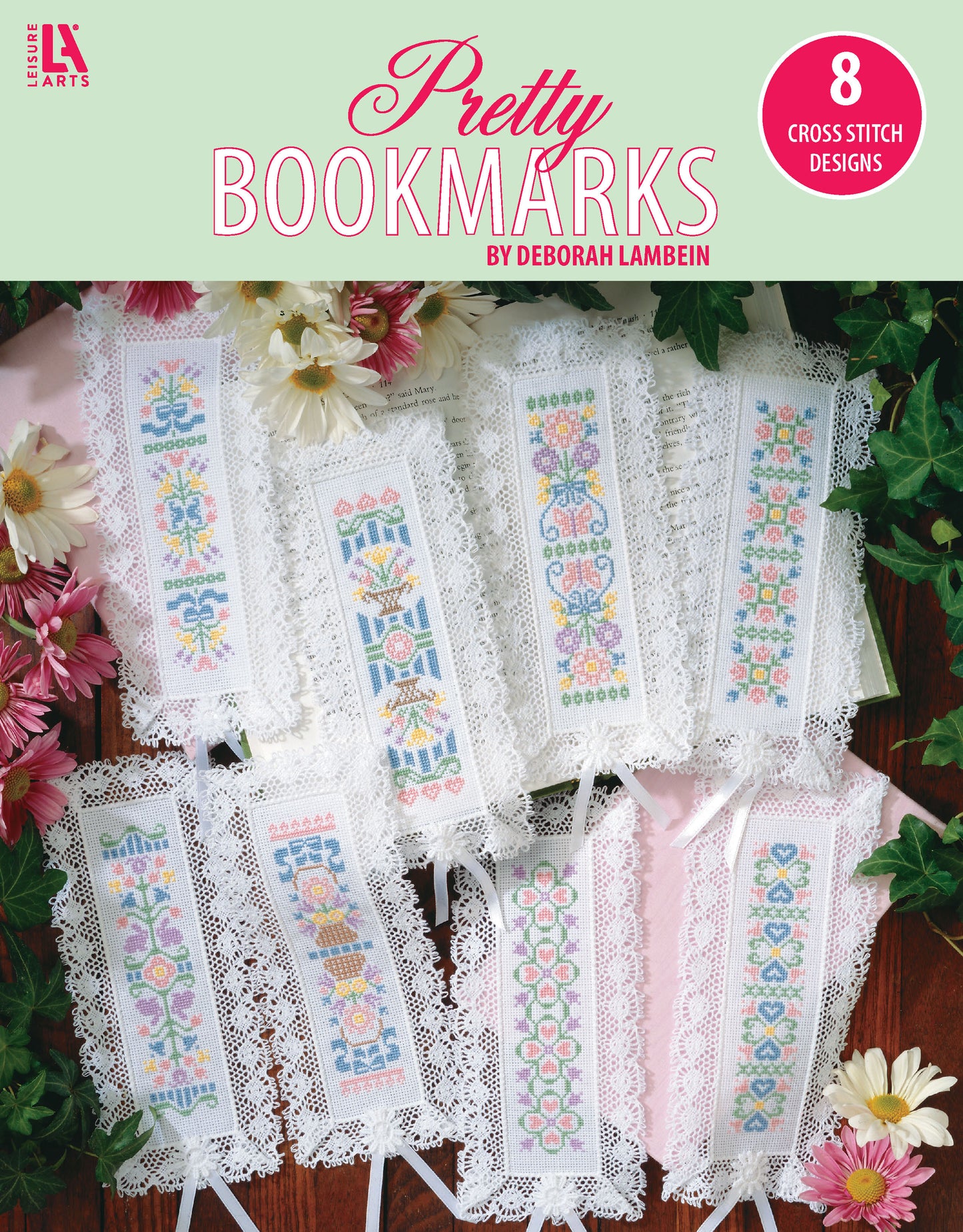 A set of 8 lace-edged bookmarks stitched with embroidery floss on 18-count aida, featuring pastel floral and geometric designs. Displayed on white cloth with pink and yellow flowers. Cross Stitch Pretty Bookmarks by Leisure Arts.