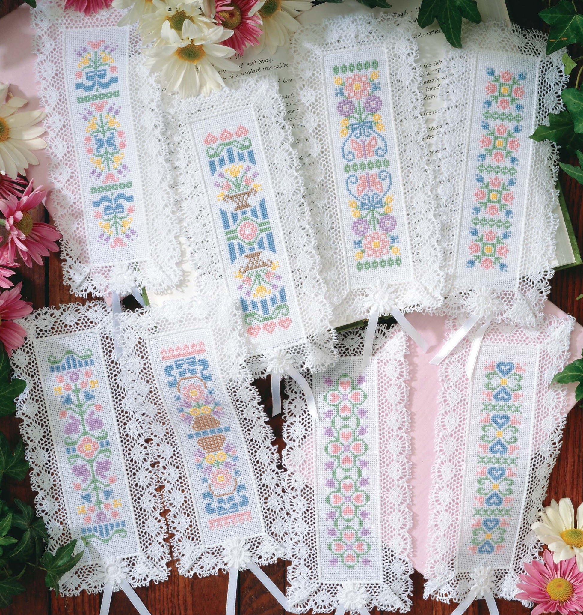 Cross Stitch Pretty Bookmarks by Leisure Arts features seven lace-edged bookmarks stitched with embroidery floss on 18 count aida, showcasing detailed floral patterns arranged among pink and white flowers on a wooden surface.