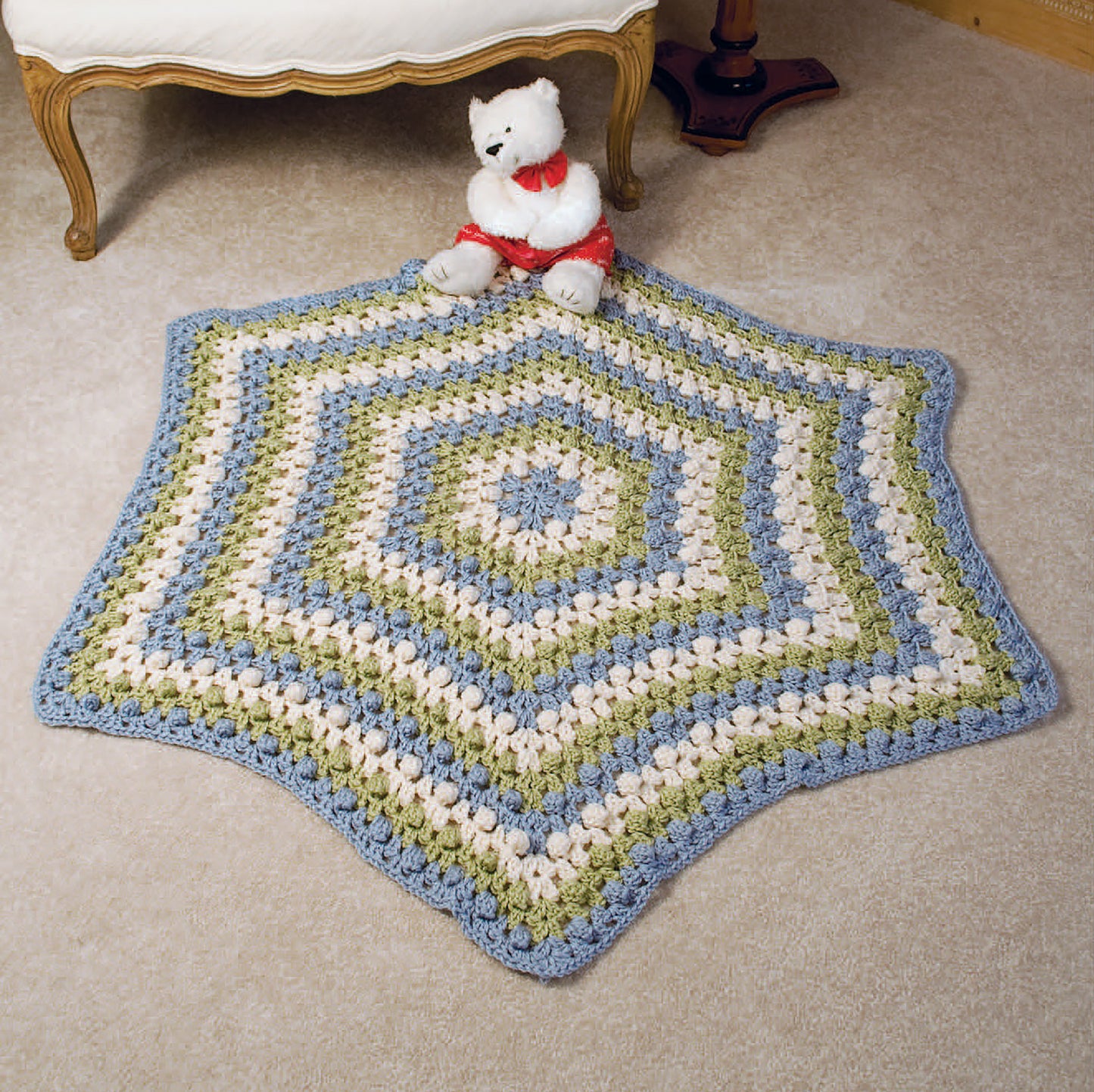 The 24-Hour Baby Afghans Digital Download from Leisure Arts features a striped, crocheted hexagonal baby blanket in blue, green, and cream displayed on a beige carpet with a white teddy bear near an upholstered chair.