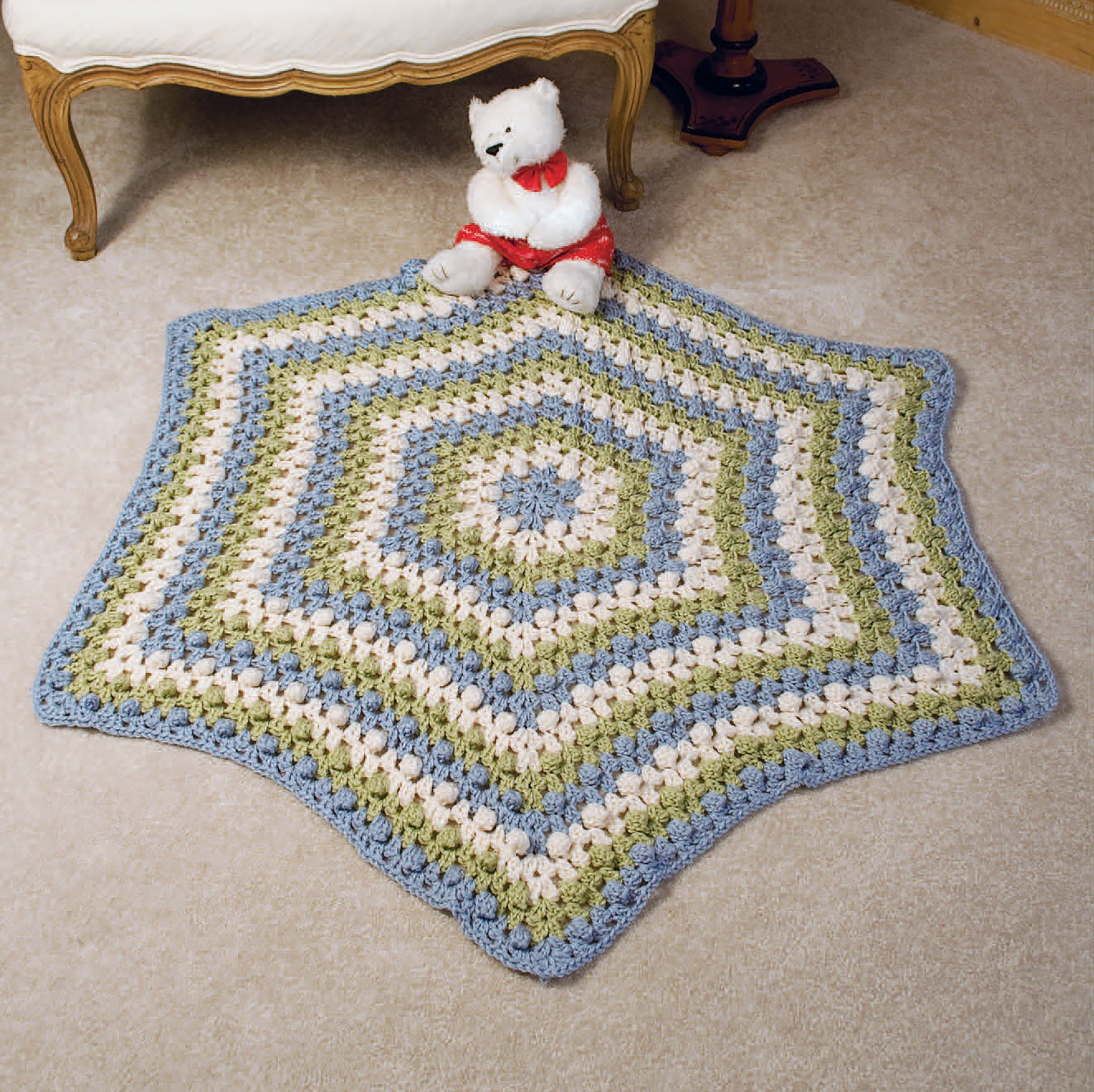 The 24-Hour Baby Afghans Digital Download from Leisure Arts features a striped, crocheted hexagonal baby blanket in blue, green, and cream displayed on a beige carpet with a white teddy bear near an upholstered chair.