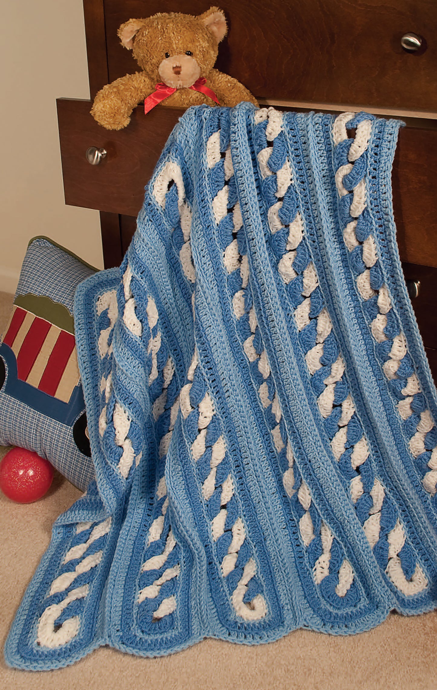 A cozy blue and white blanket, made with the Leisure Arts 24-Hour Baby Afghans Digital Download pattern, hangs from an open dresser drawer beside a brown teddy bear. Nearby are a blue pillow featuring a train design and a red ball on the carpet.