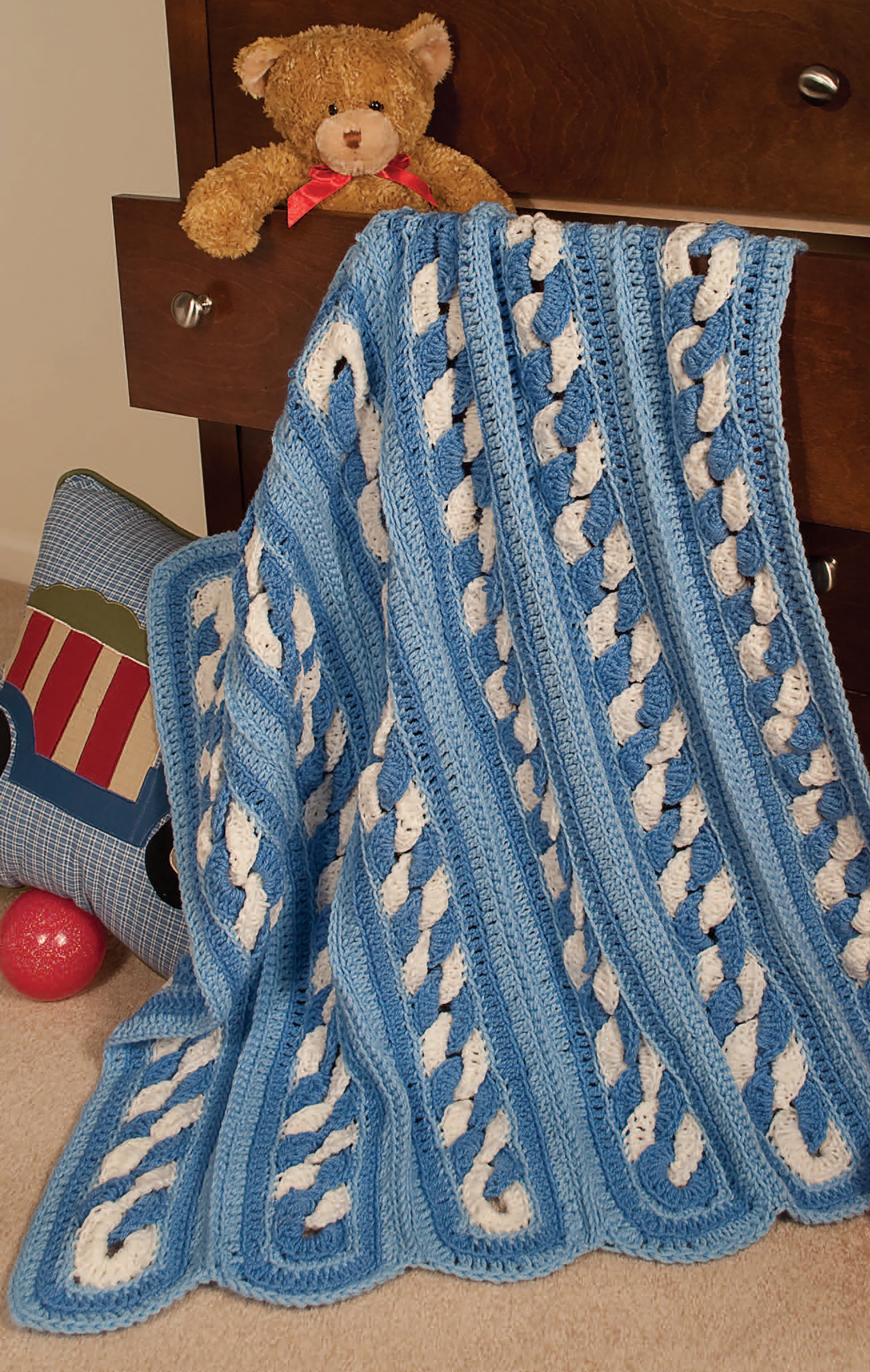 A cozy blue and white blanket, made with the Leisure Arts 24-Hour Baby Afghans Digital Download pattern, hangs from an open dresser drawer beside a brown teddy bear. Nearby are a blue pillow featuring a train design and a red ball on the carpet.