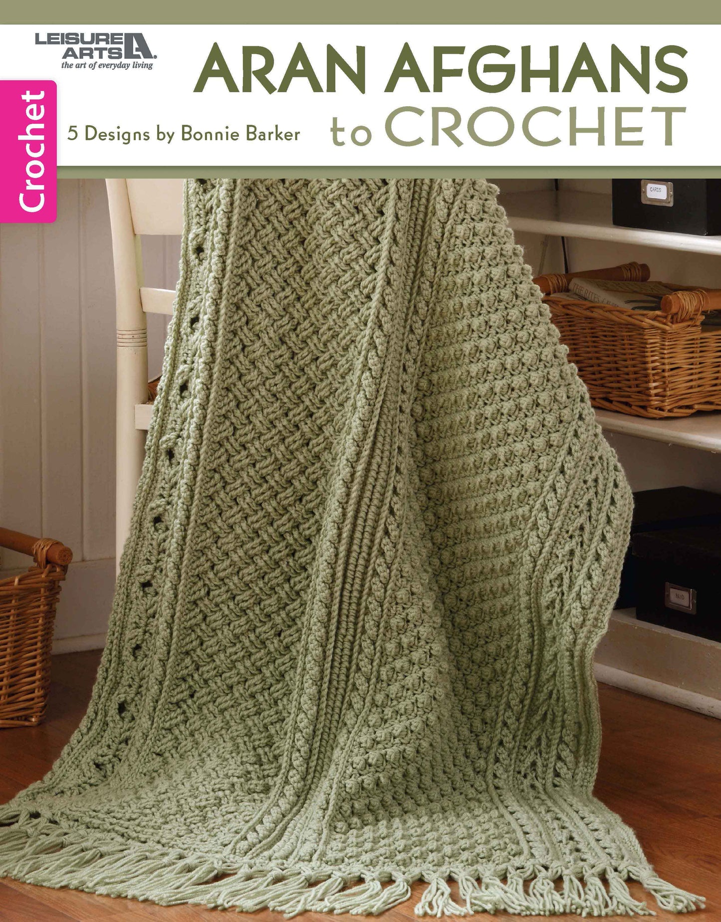 A light green, textured afghan made with medium weight yarn and tassels drapes over a chair. The book "Aran Afghans to Crochet" by Leisure Arts offers 5 intricate intermediate crochet patterns by Bonnie Barker.