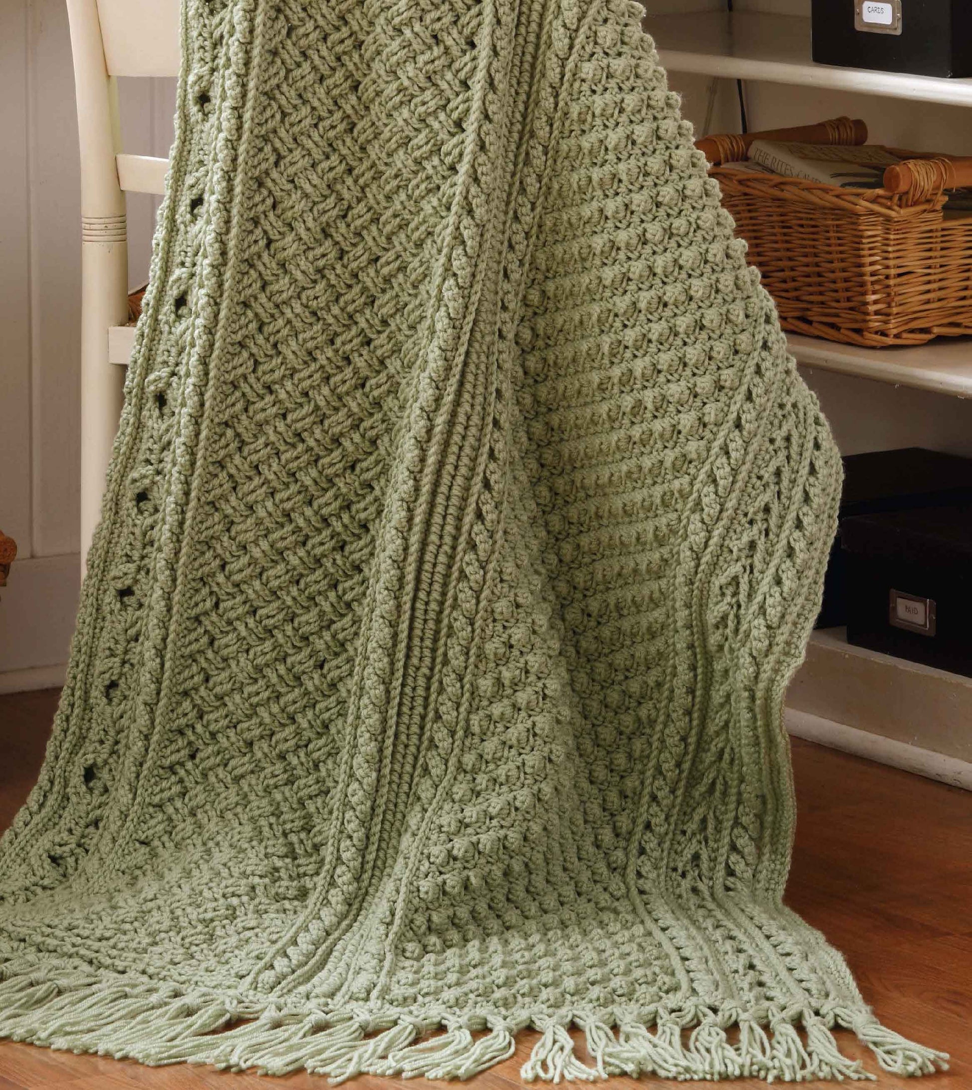 The "Aran Afghans to Crochet" book by Leisure Arts showcases medium-weight, textured knit throws in light green, as seen draped over a chair in a cozy room with shelves and wicker baskets in the background.