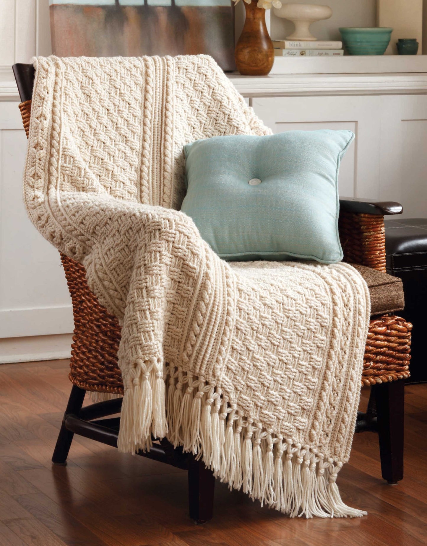 A cozy room features a woven armchair with an Aran Afghans to Crochet throw by Leisure Arts draped over it, complemented by a light blue cushion on a wooden floor, with white cabinetry and decor in the background.