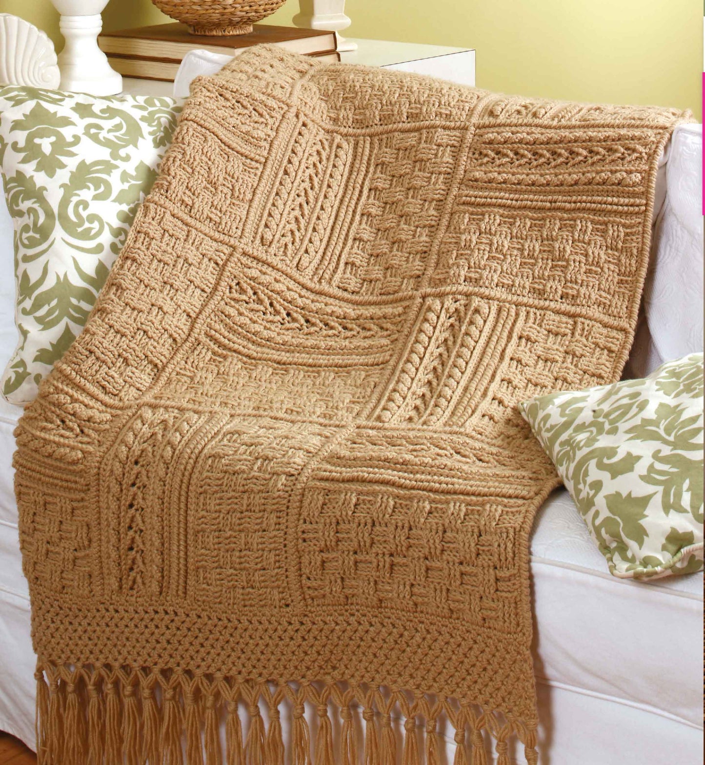 The "Aran Afghans to Crochet" by Leisure Arts, a tan, hand-knitted afghan with textured square patterns and fringe, is draped over a white couch accented with green and white floral pillows.