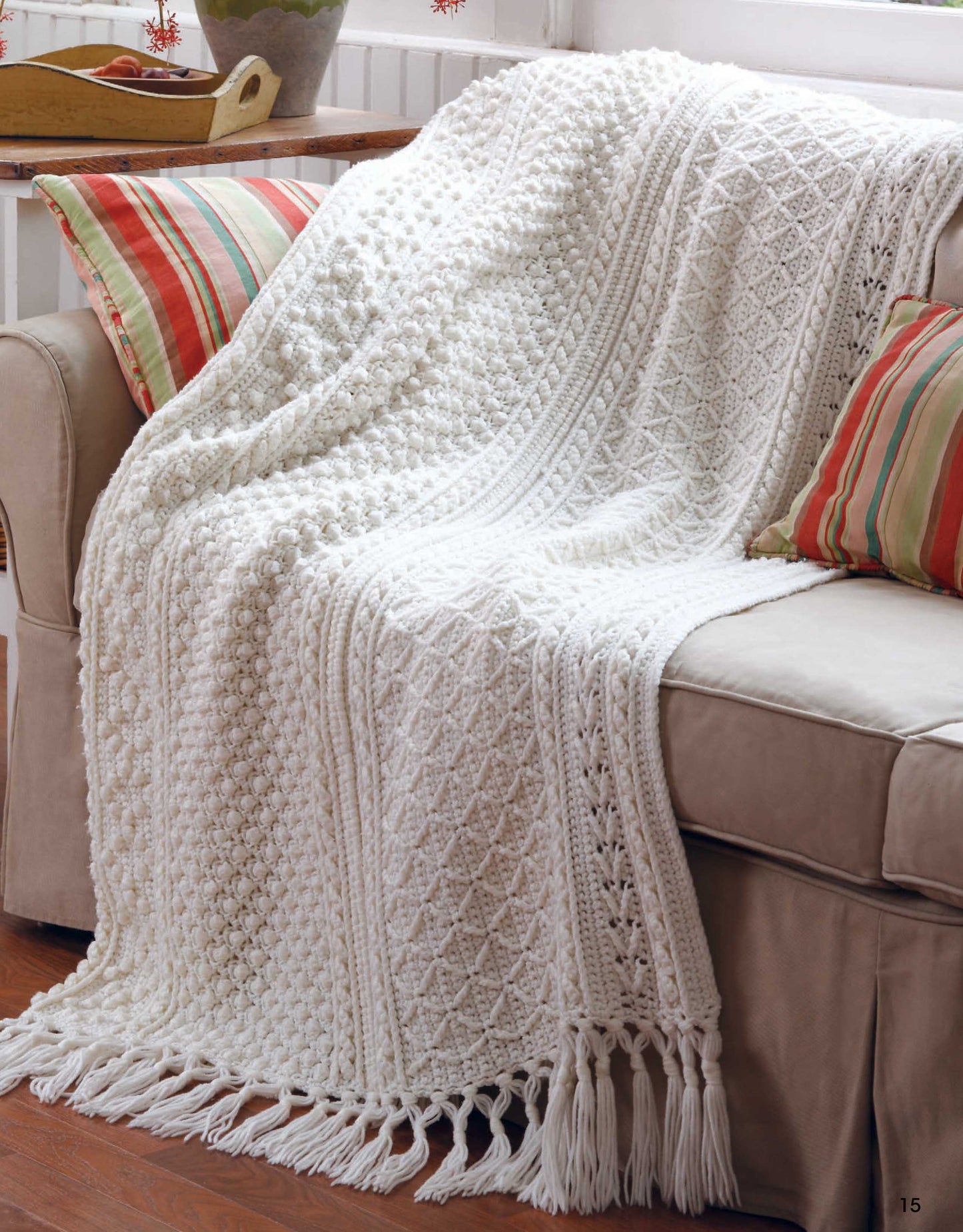 A beige sofa with striped red and green pillows by a window is draped with a white, textured knit blanket inspired by Leisure Arts' "Aran Afghans to Crochet.