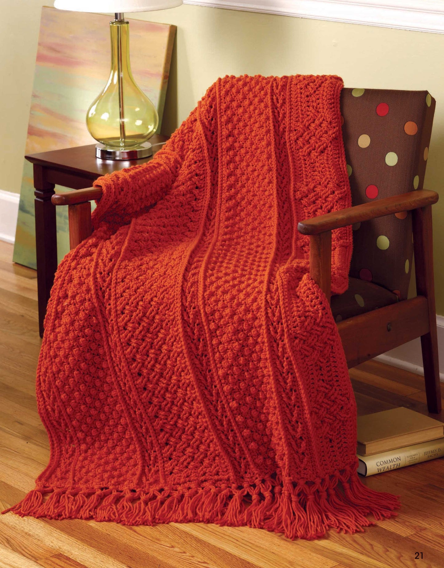 A cozy knit blanket from the "Aran Afghans to Crochet" collection by Leisure Arts drapes over a polka dot brown armchair, with a sleek glass lamp, abstract art, and wooden flooring completing the modern setting.