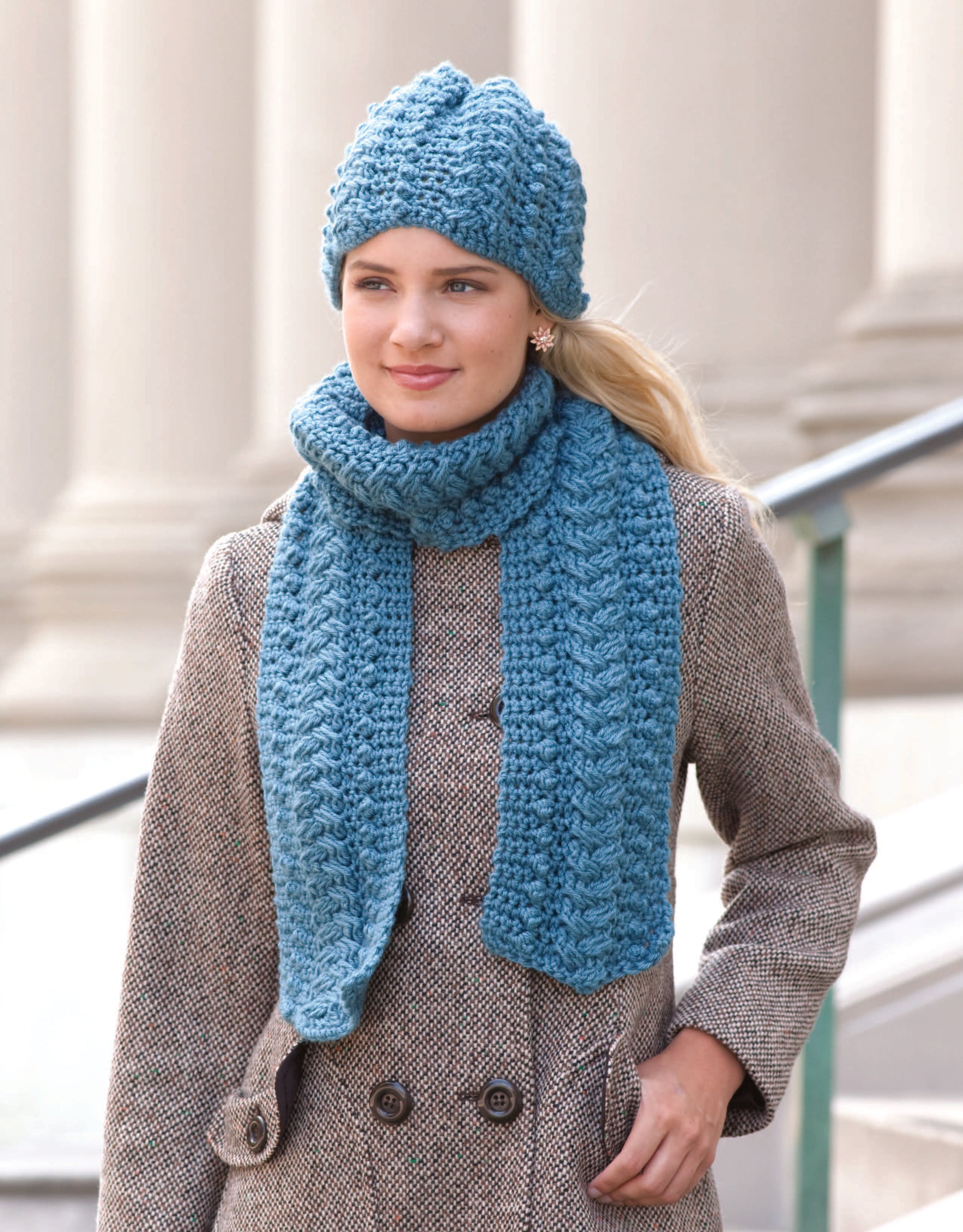 A woman stands outdoors before stone columns, wearing a light brown coat and the chunky blue Noggins And Necks set by Leisure Arts. Her look, featuring a cozy hat and scarf, offers perfect inspiration for intermediate crochet projects.