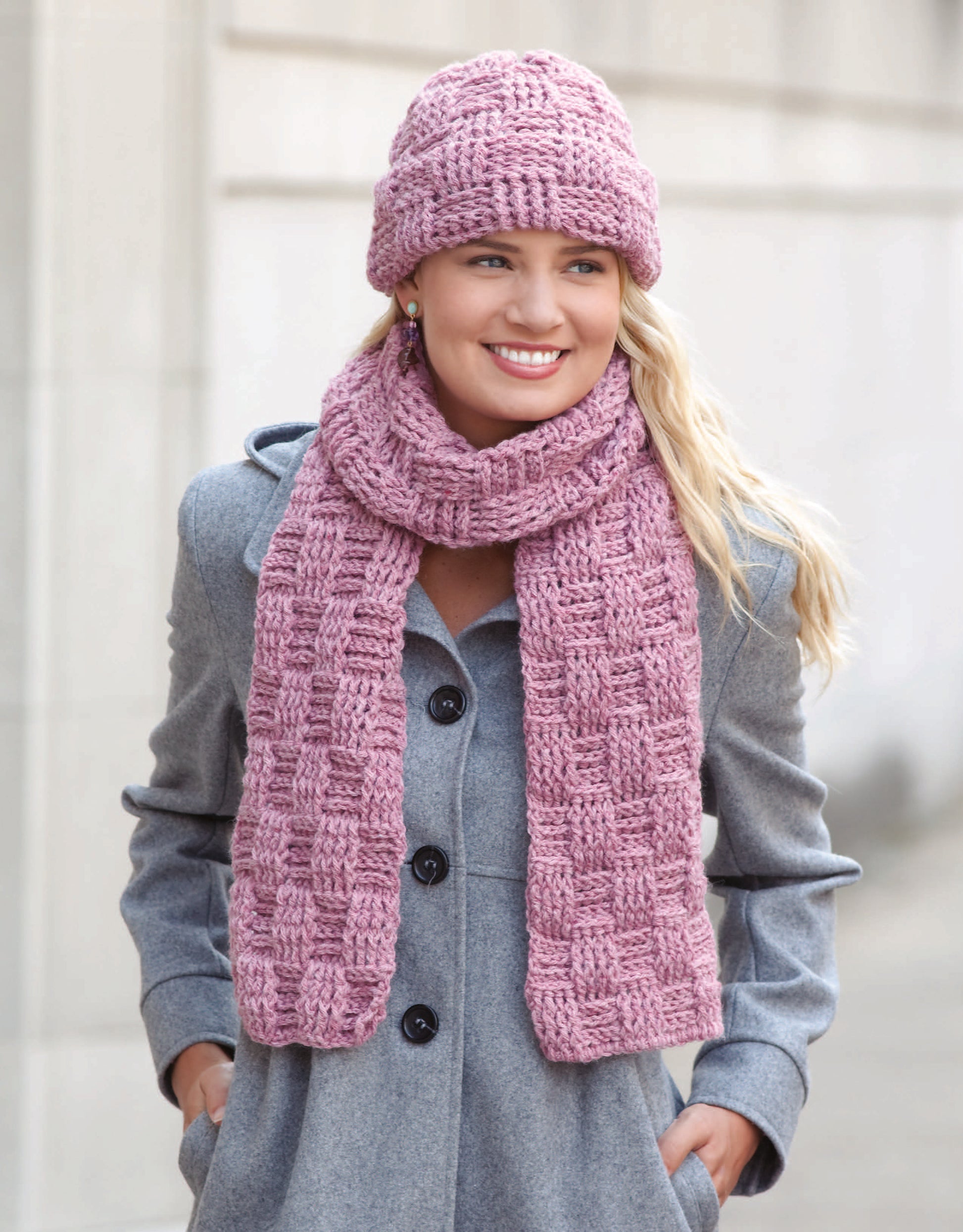 A woman stands outdoors, smiling, in a light gray coat and a pink crocheted hat and scarf made from the Noggins And Necks Digital Download by Leisure Arts. The background is a softly blurred stone wall.