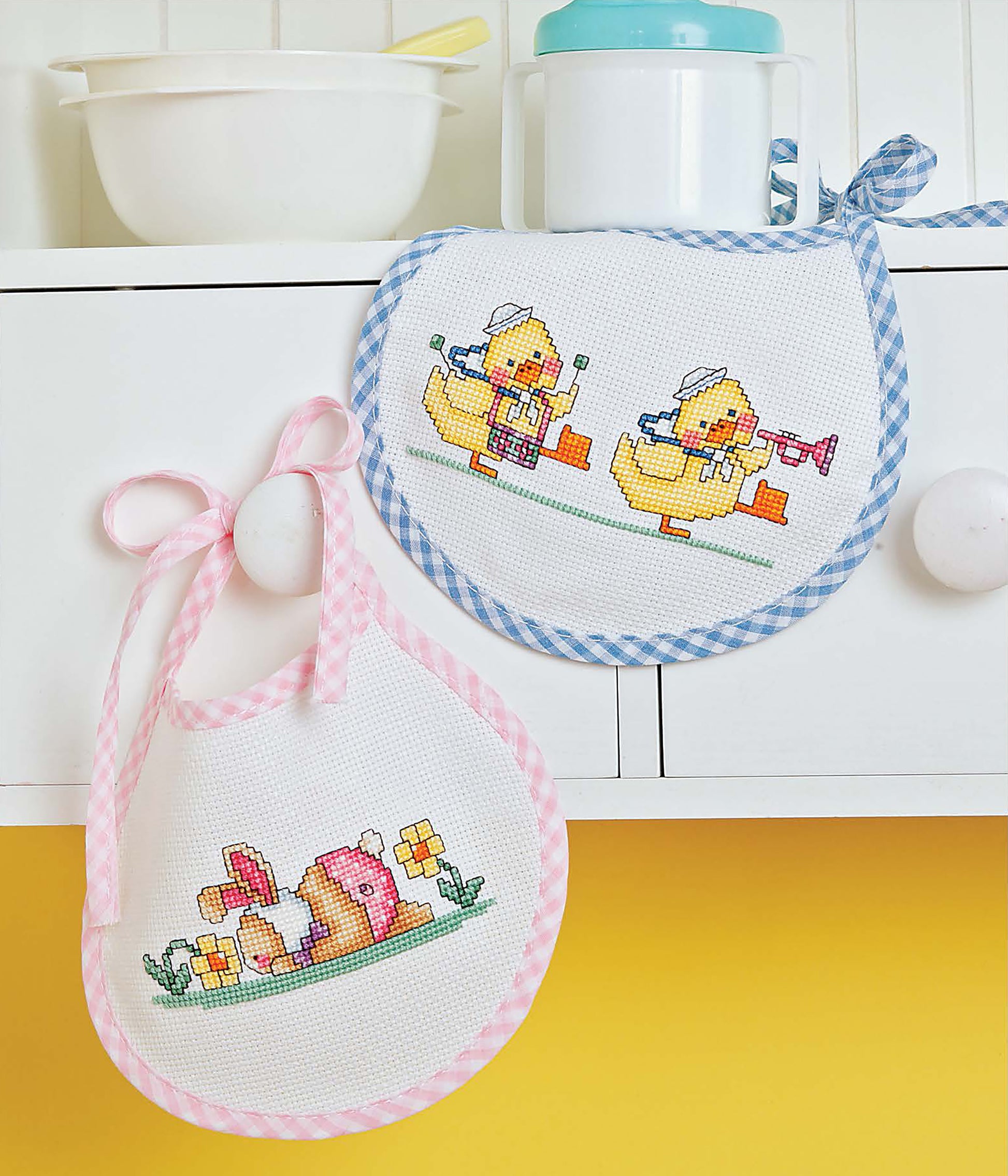 Two baby bibs from "The Best of Cross Stitch Basics" by Leisure Arts hang on a white cabinet, showcasing beginner-friendly cross stitch patterns with ducks, a bunny, and flowers—ideal for handmade home decor or embroidery projects.