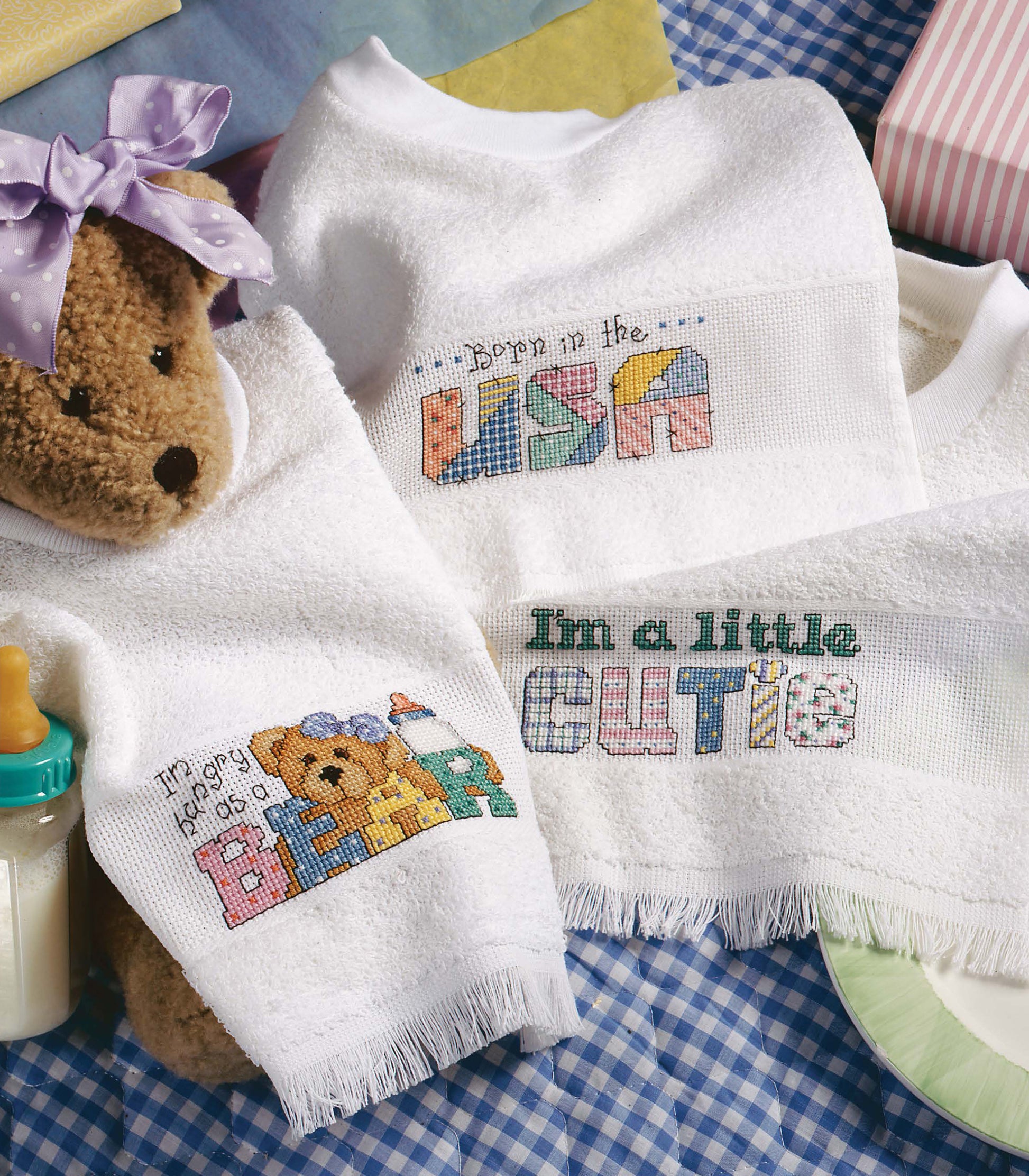Leisure Arts' "The Best of Cross Stitch Basics" features three white baby bibs with playful sayings—“Born in the USA,” “I’m a guy so do as I BEAR,” and “I’m a little CUTIE”—ideal for beginner embroidery and home decor crafts.