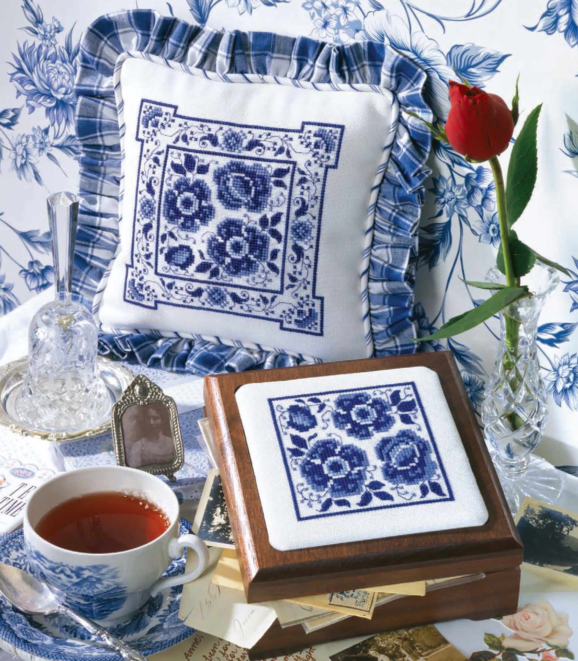 Create a charming scene featuring "The Best of Cross Stitch Basics" by Leisure Arts, with a blue and white cross-stitched pillow and box, red rose in vase, teacup, vintage letters, photo frame, crystal decanter, and blue floral wallpaper.