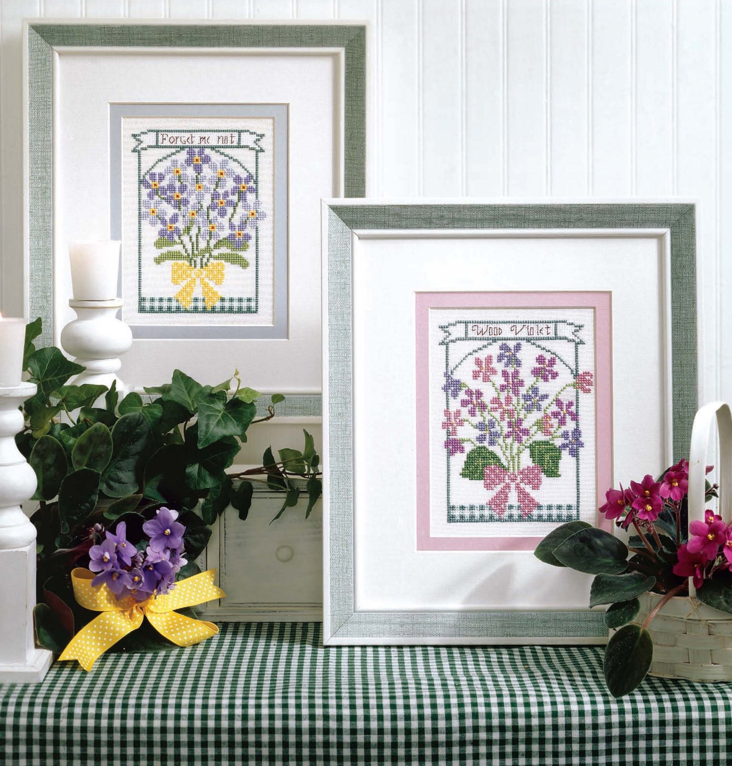 Two framed floral cross-stitch artworks, made with beginner patterns from "The Best of Cross Stitch Basics" by Leisure Arts, sit on a green checkered tablecloth among potted plants and baskets for cozy cottage-style home decor.