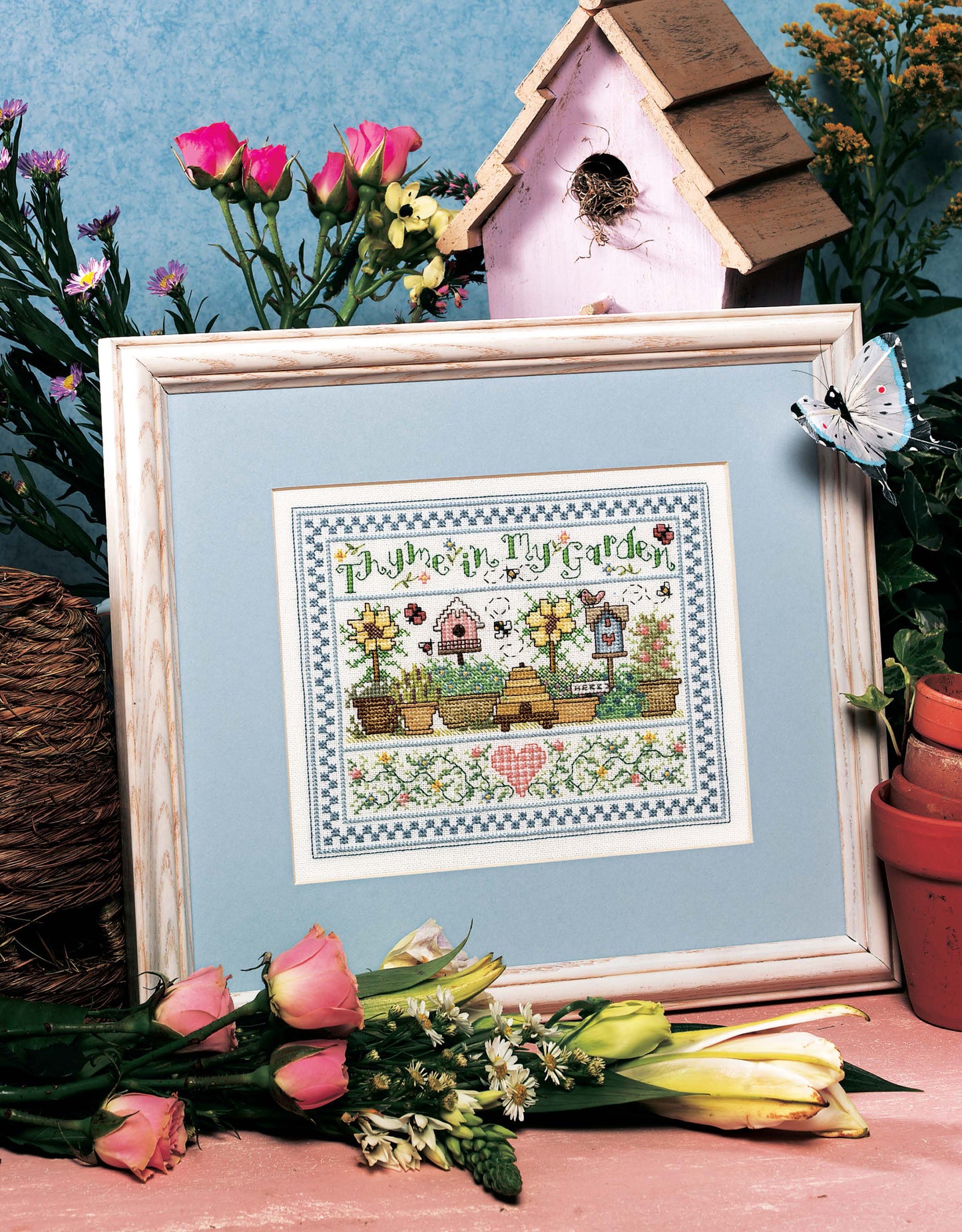 The Best of Cross Stitch Basics by Leisure Arts features a framed Thyme in Thy Garden design displayed with flowers, a birdhouse, butterfly, and twine—a charming scene ideal for home decor or beginner embroidery.