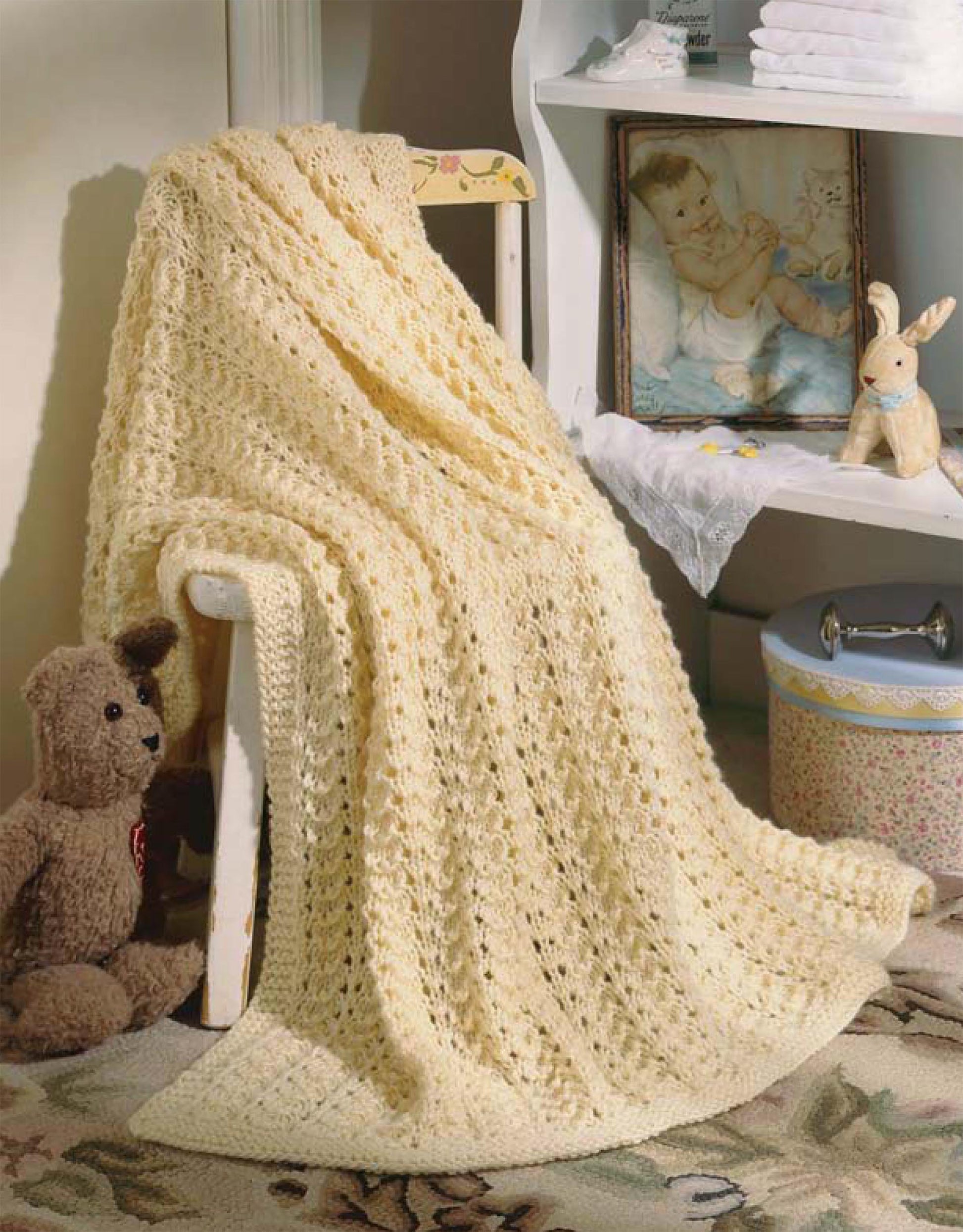 A pale yellow blanket from "Our Best Knit Baby Afghans, Book 2" by Leisure Arts is draped over a chair beside a brown teddy bear, a blue container, and shelves with a framed baby photo, stuffed rabbit, and stacked white towels.