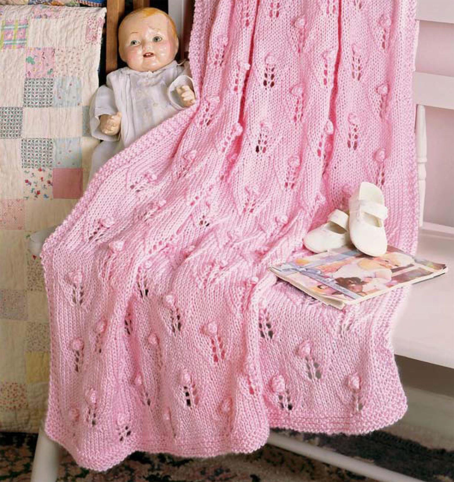 A vintage doll sits on a white chair draped with a pink blanket made from patterns in "Our Best Knit Baby Afghans, Book 2" by Leisure Arts. Nearby, a quilt, baby shoes, and a children’s book create a nostalgic, cozy feel.