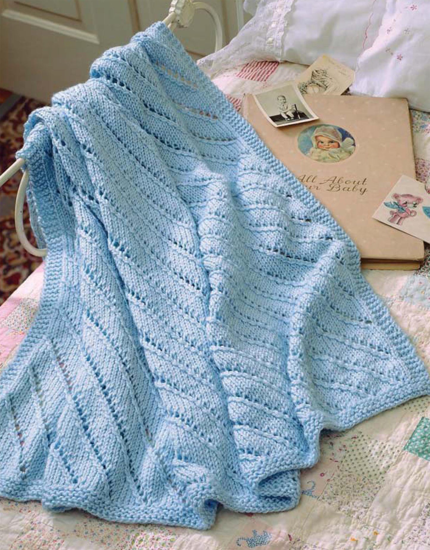 A light blue knit blanket, made with circular needles, hangs over a white bed rail. On the bed are vintage photos, a pastel patterned quilt, and Leisure Arts' "Our Best Knit Baby Afghans, Book 2.