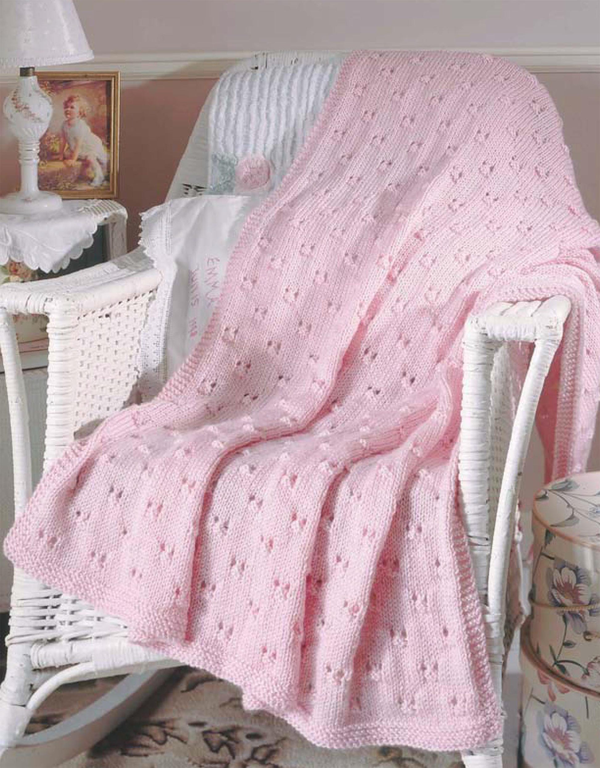 A pink blanket made with patterns from Leisure Arts' "Our Best Knit Baby Afghans, Book 2" is draped over a white wicker chair in a cozy room, with a cushion, side table, and framed photo in the background.