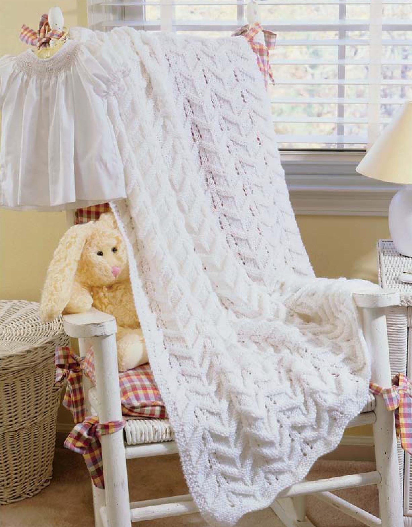 A cozy white blanket inspired by patterns from "Our Best Knit Baby Afghans, Book 2" by Leisure Arts is draped over a rocking chair with plaid cushions, a yellow plush bunny, and a baby dress. Soft light filters in through window blinds behind.