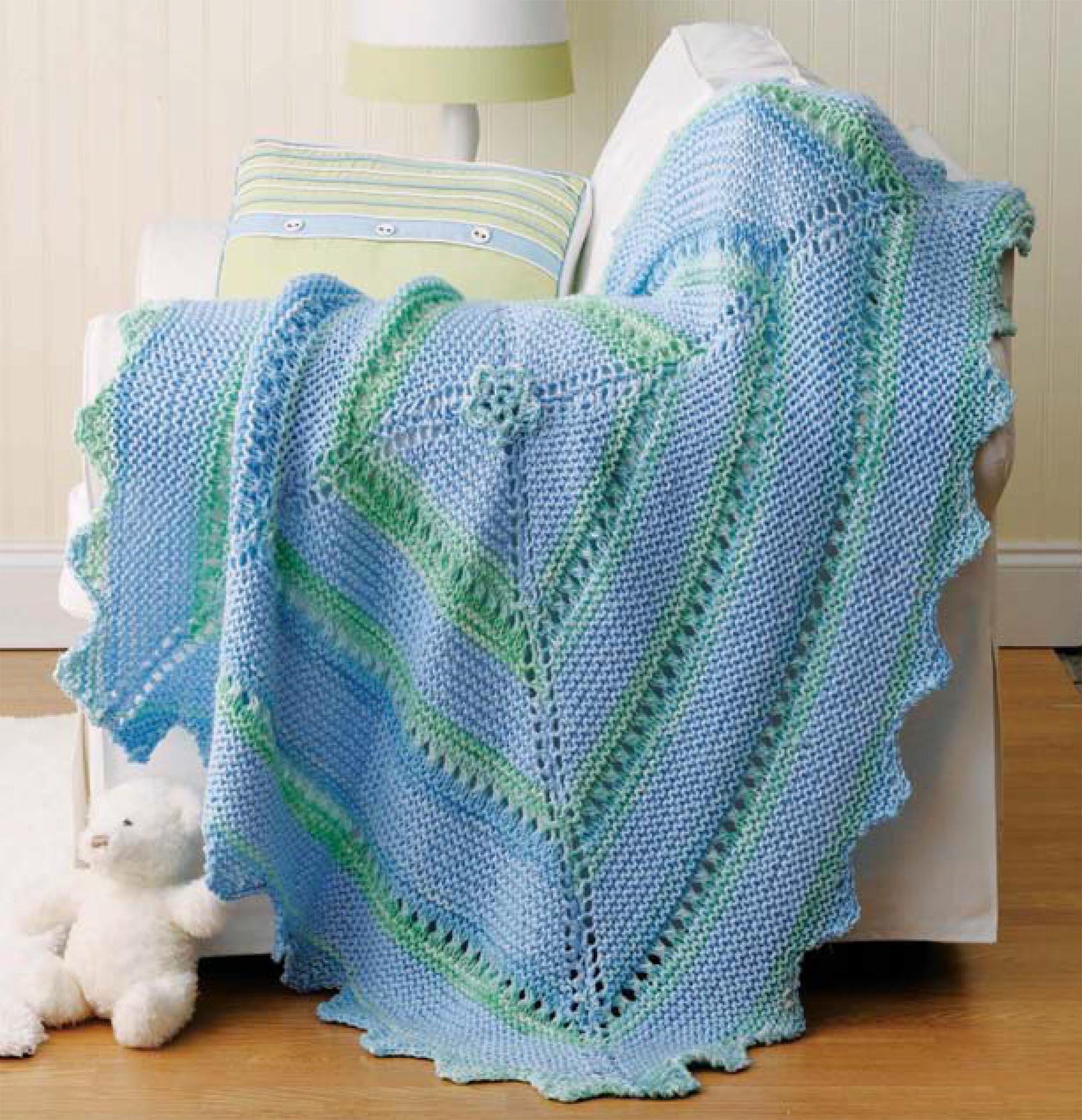The Leisure Arts "Our Best Knit Baby Afghans, Book 2" features a light blue and green lace-patterned blanket knitted with circular needles, displayed on a white armchair beside a pastel pillow and lamp, with a white teddy bear on the floor.