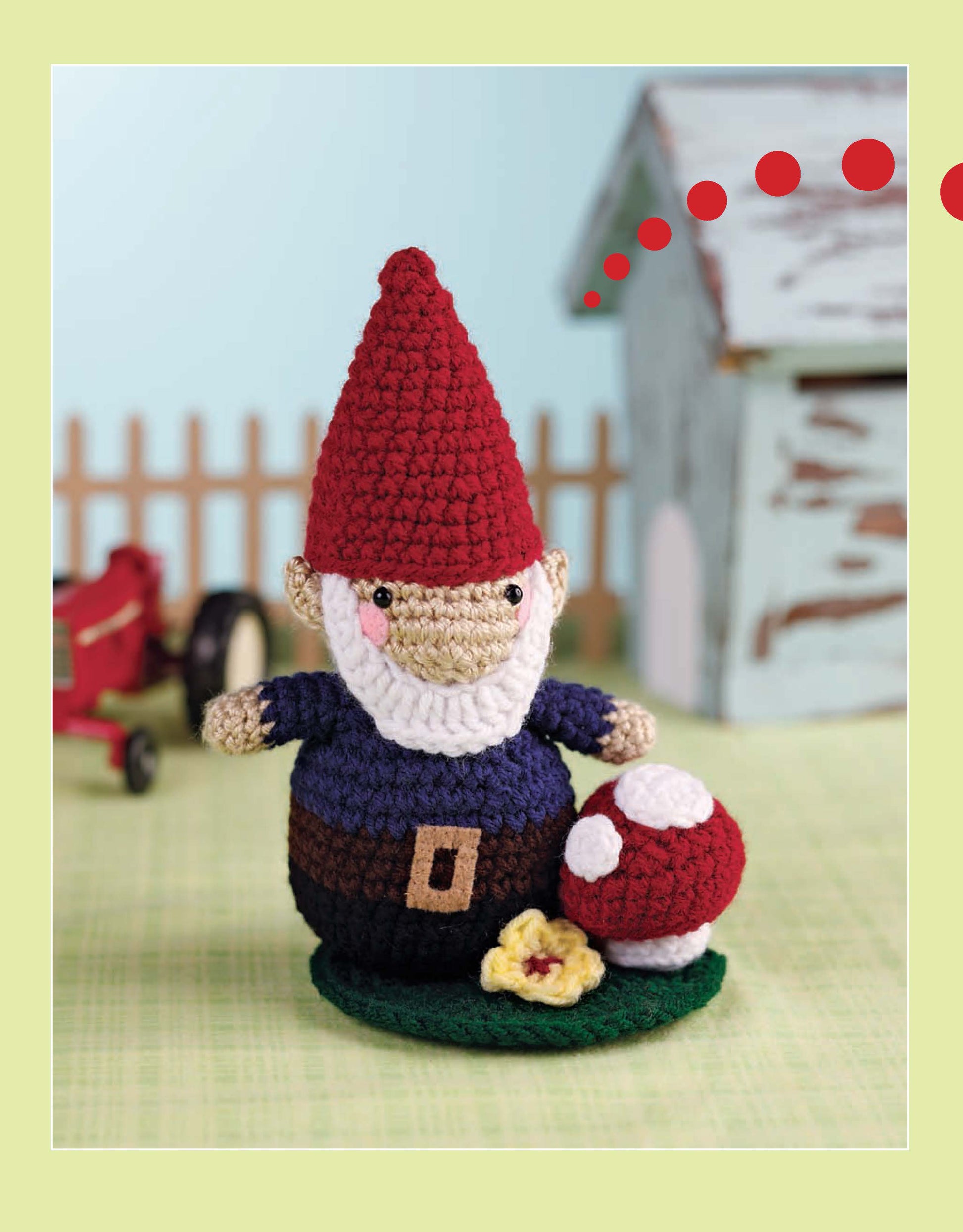 A crocheted gnome from Leisure Arts’ "More Cute Little Animals To Crochet" stands on green grass beside a mushroom—perfect for amigurumi or beginner crochet inspiration. A toy house and red tractor blur in the background.