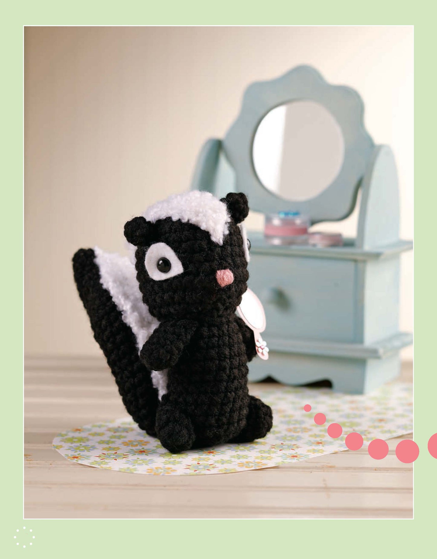 A black and white crocheted skunk plush from "More Cute Little Animals To Crochet" by Leisure Arts stands before a small blue vanity mirror with pastel accessories on a light floral mat.