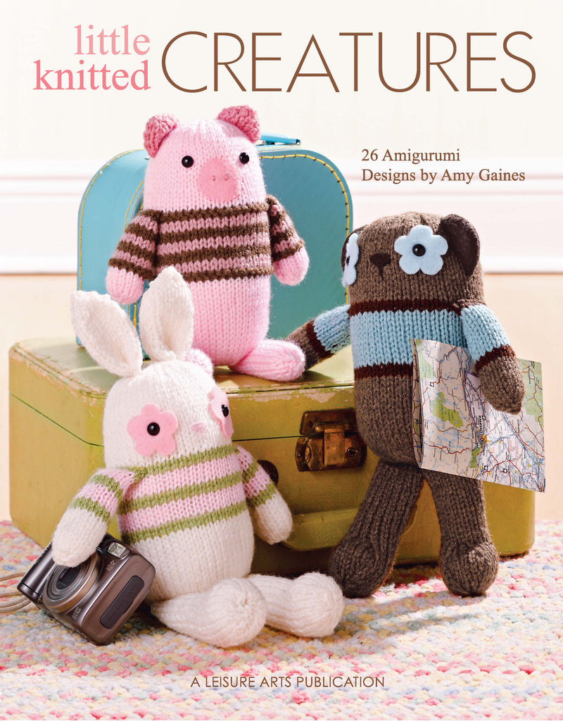 Three knitted animals—a pink pig, white bunny, and brown bear—sit by a yellow suitcase. The bear holds a map, the bunny has a camera. Above: Little Knitted Creatures by Leisure Arts, perfect for beginner knitting projects. Design credits included.